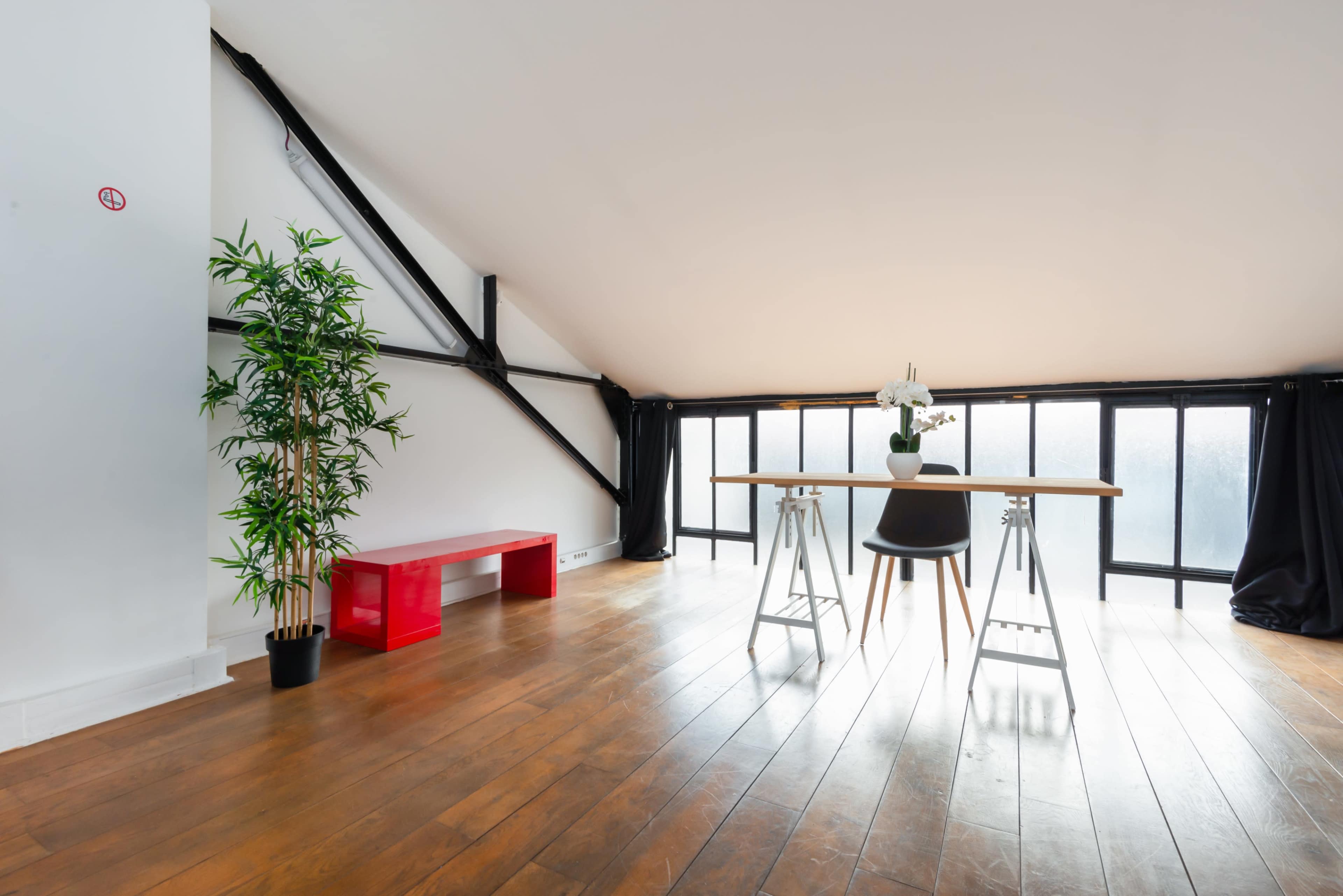 The image shows a bright, minimalist loft space with a wooden floor, a simple dining table, a plant, and large windows.