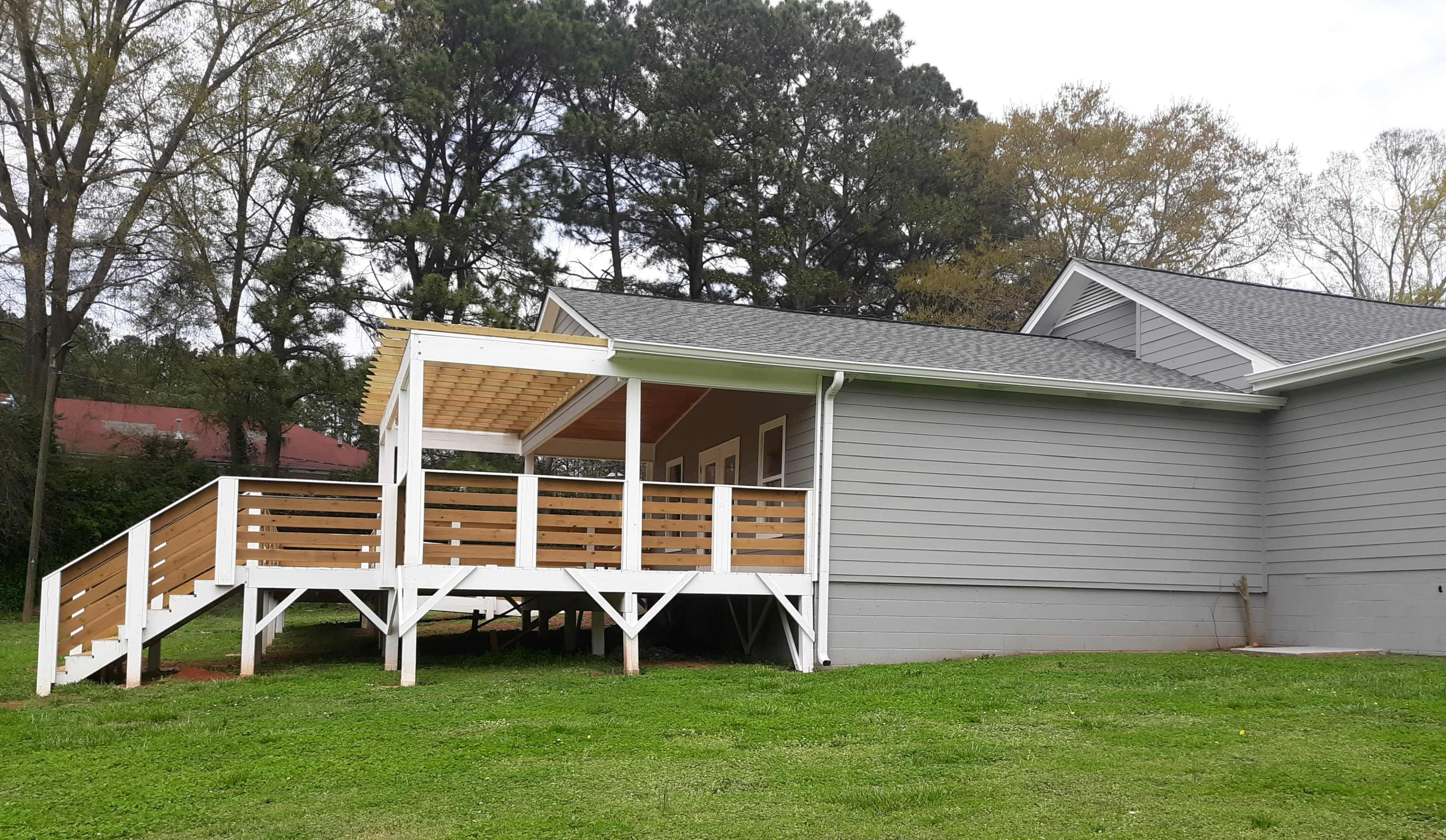 The image shows a gray house with a wooden deck extending from its side, surrounded by trees and grass.