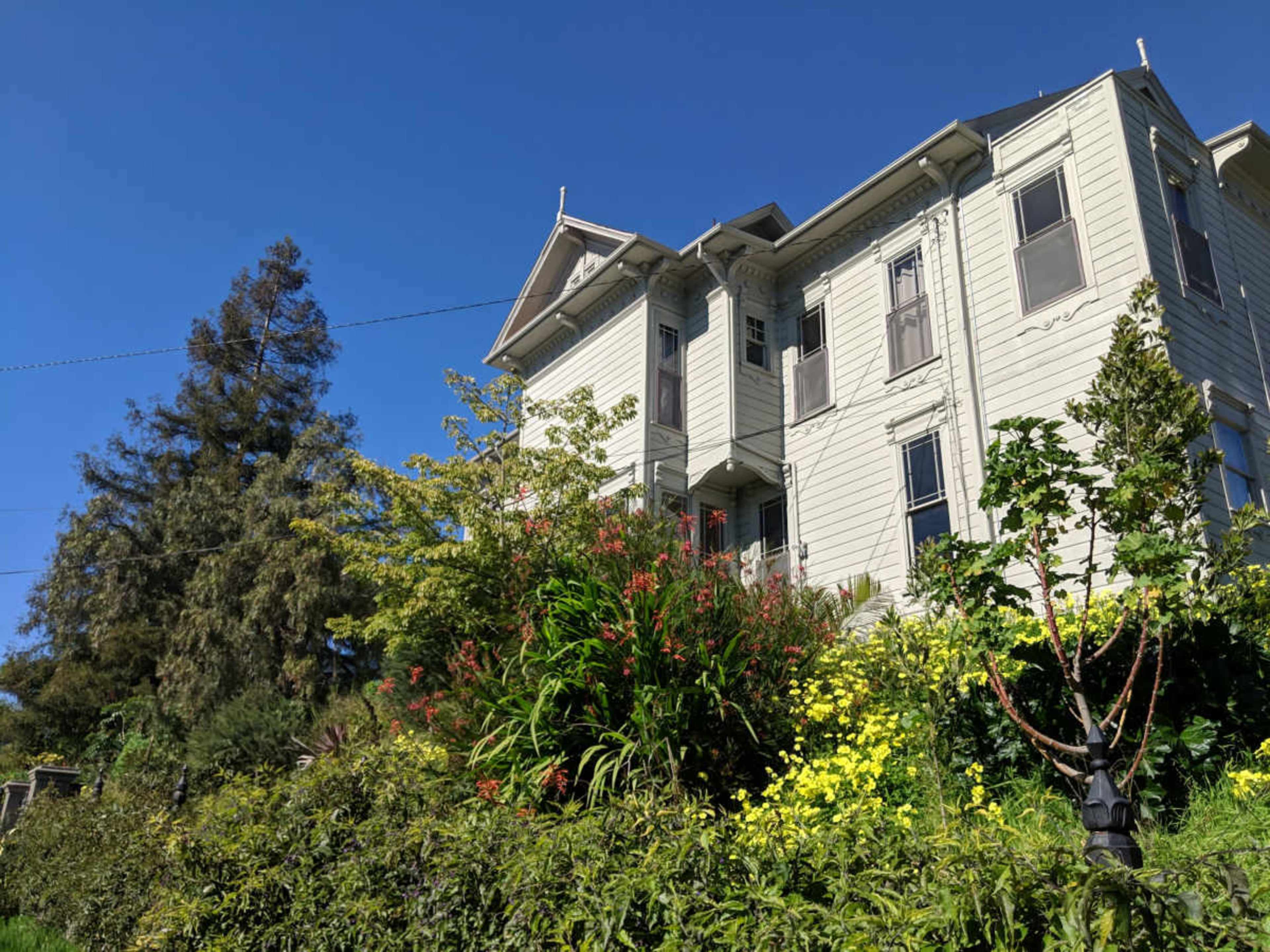 A three-story Victorian house is surrounded by lush greenery and colorful flowers under a clear blue sky.
