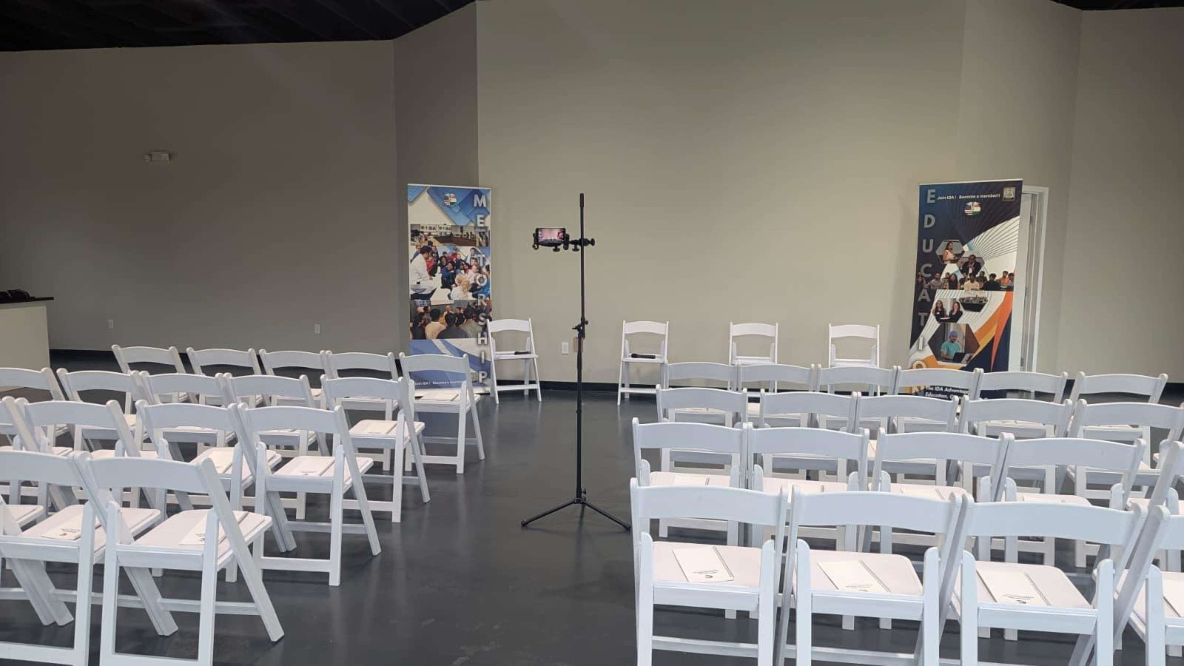 The image shows a meeting room set up with white folding chairs arranged in rows, a mobile stand in the center, and two banners on either side.