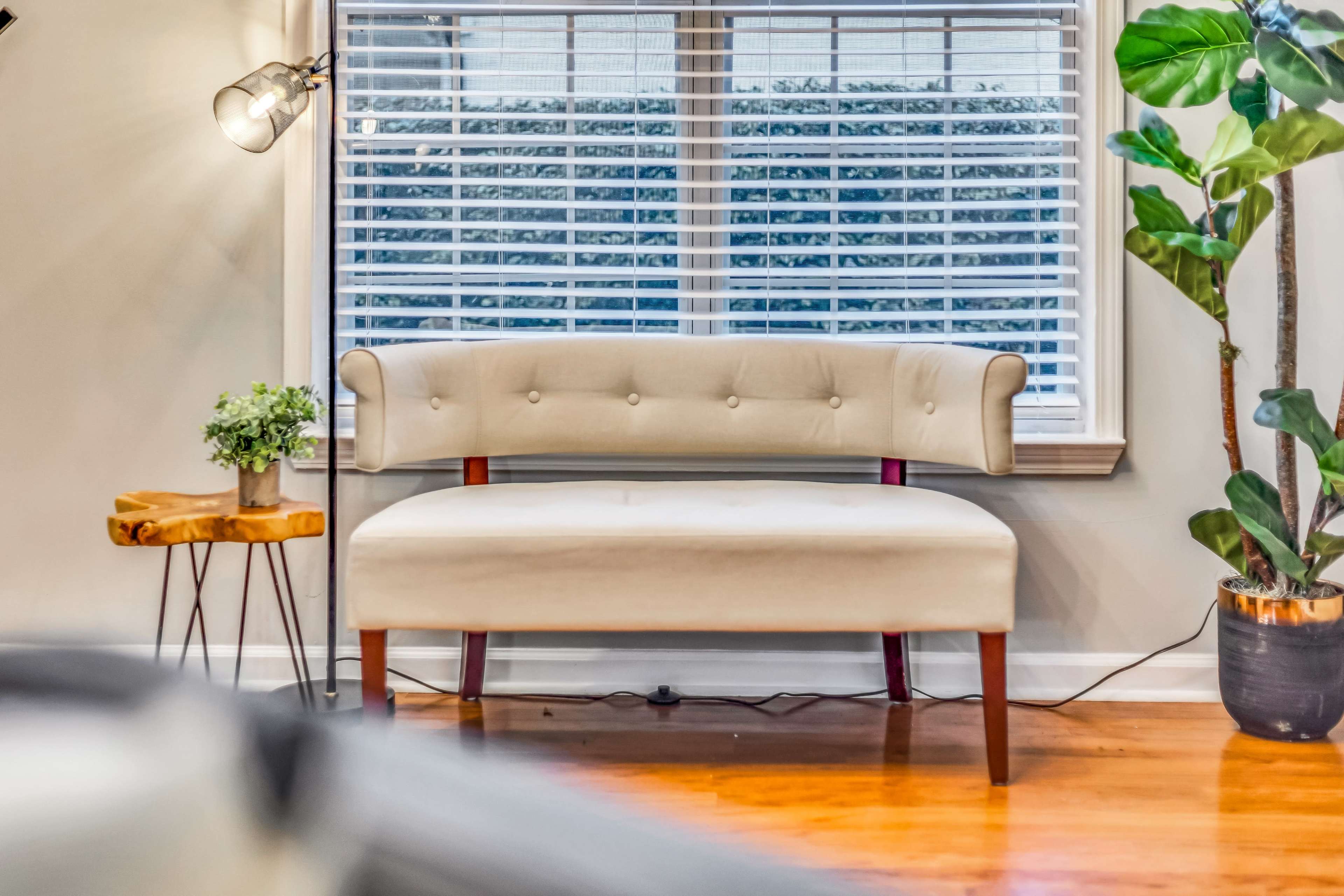 A light-colored upholstered bench is positioned by a window with horizontal blinds, accompanied by a side table and a potted plant.