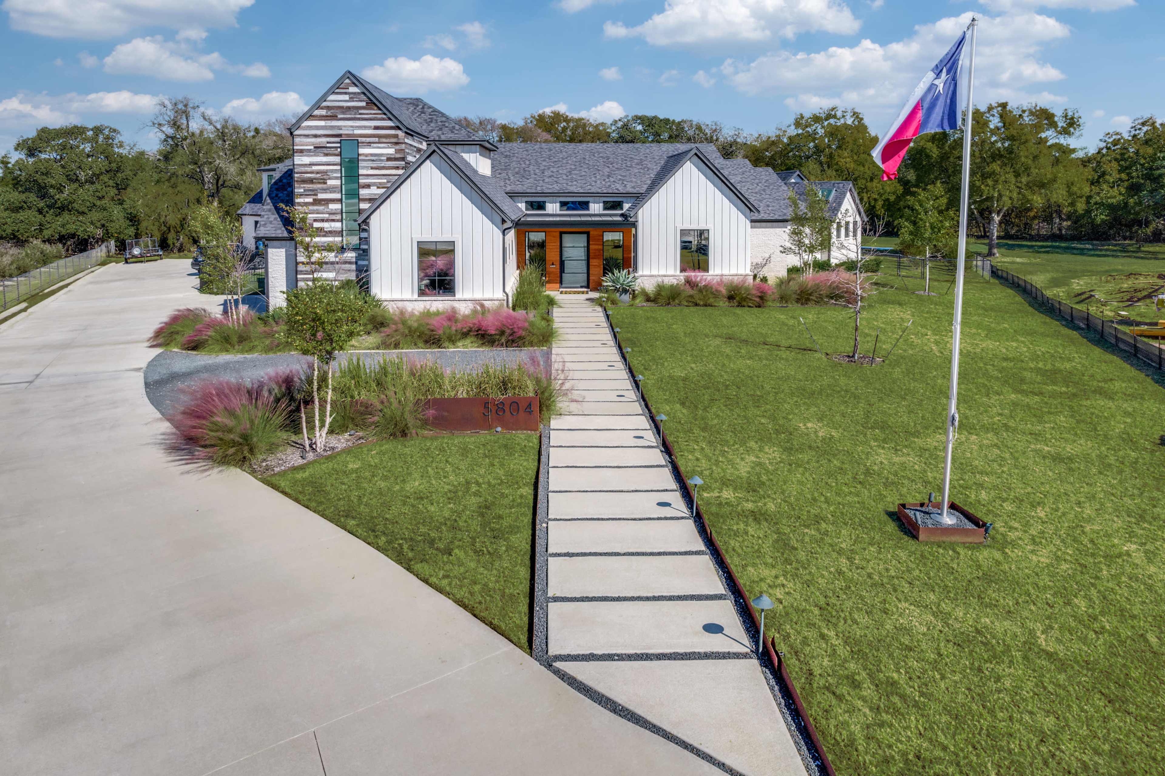 A modern house with a mix of white and gray siding, featuring large front windows and a Texas flag, is set against a landscaped lawn with a concrete path leading to the entrance.