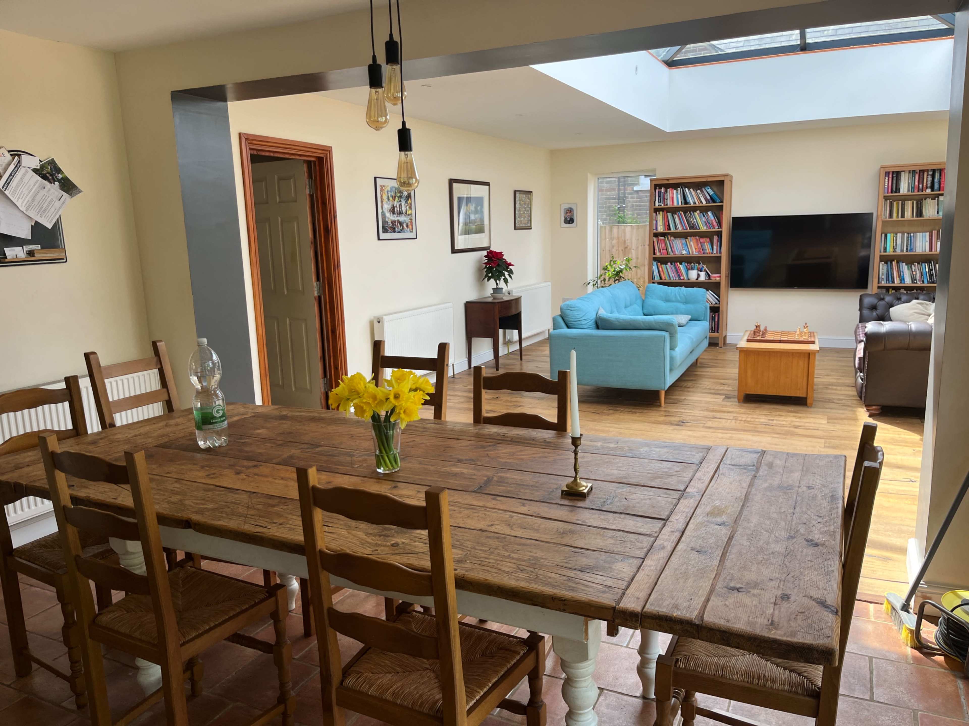 The image shows a spacious room featuring a wooden dining table with chairs, a well-lit area with a sofa and bookshelves in the background, and a large window allowing natural light.