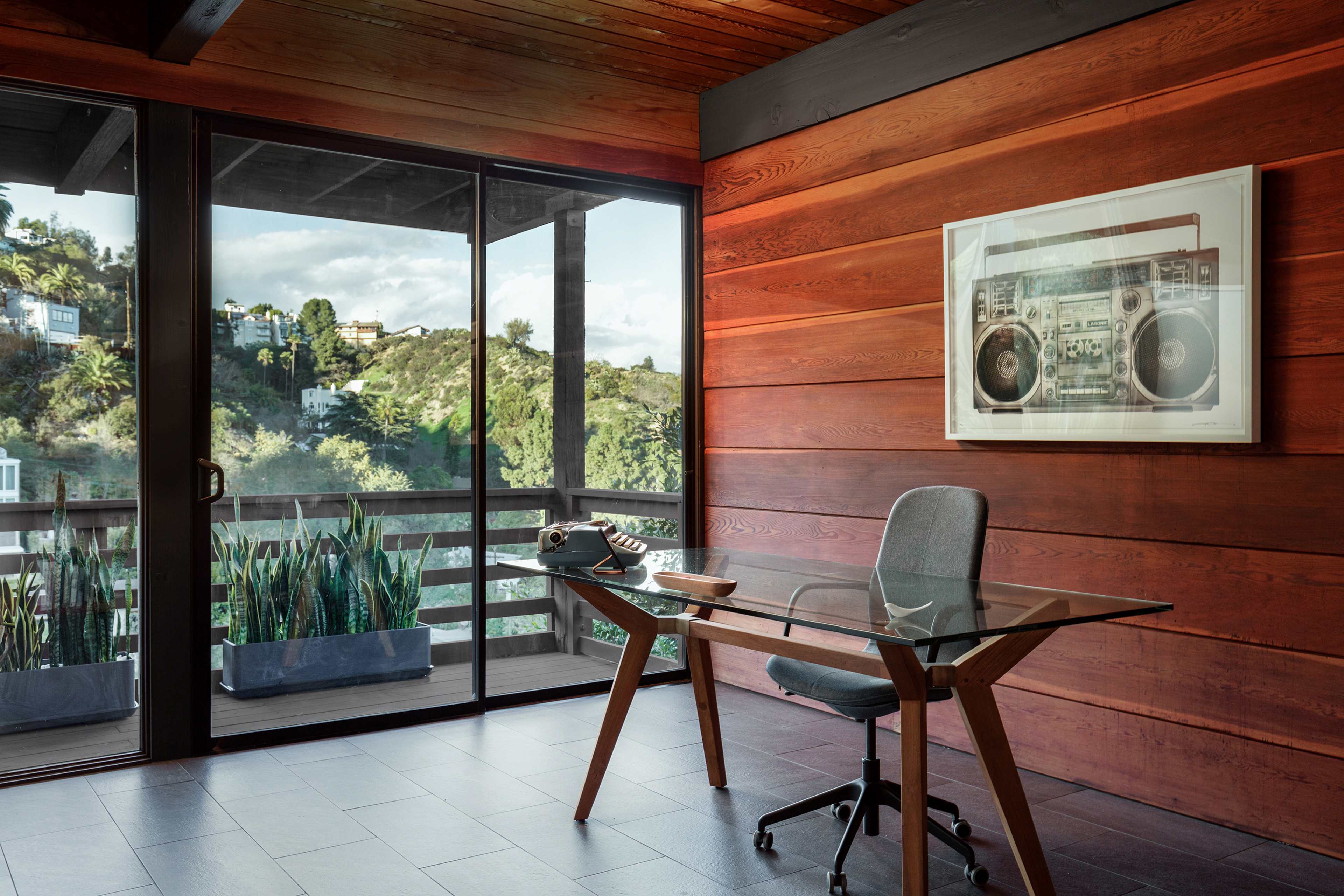 A modern office features a glass desk and a chair, positioned beside a large window with a view of green hillside, framed by wooden walls.