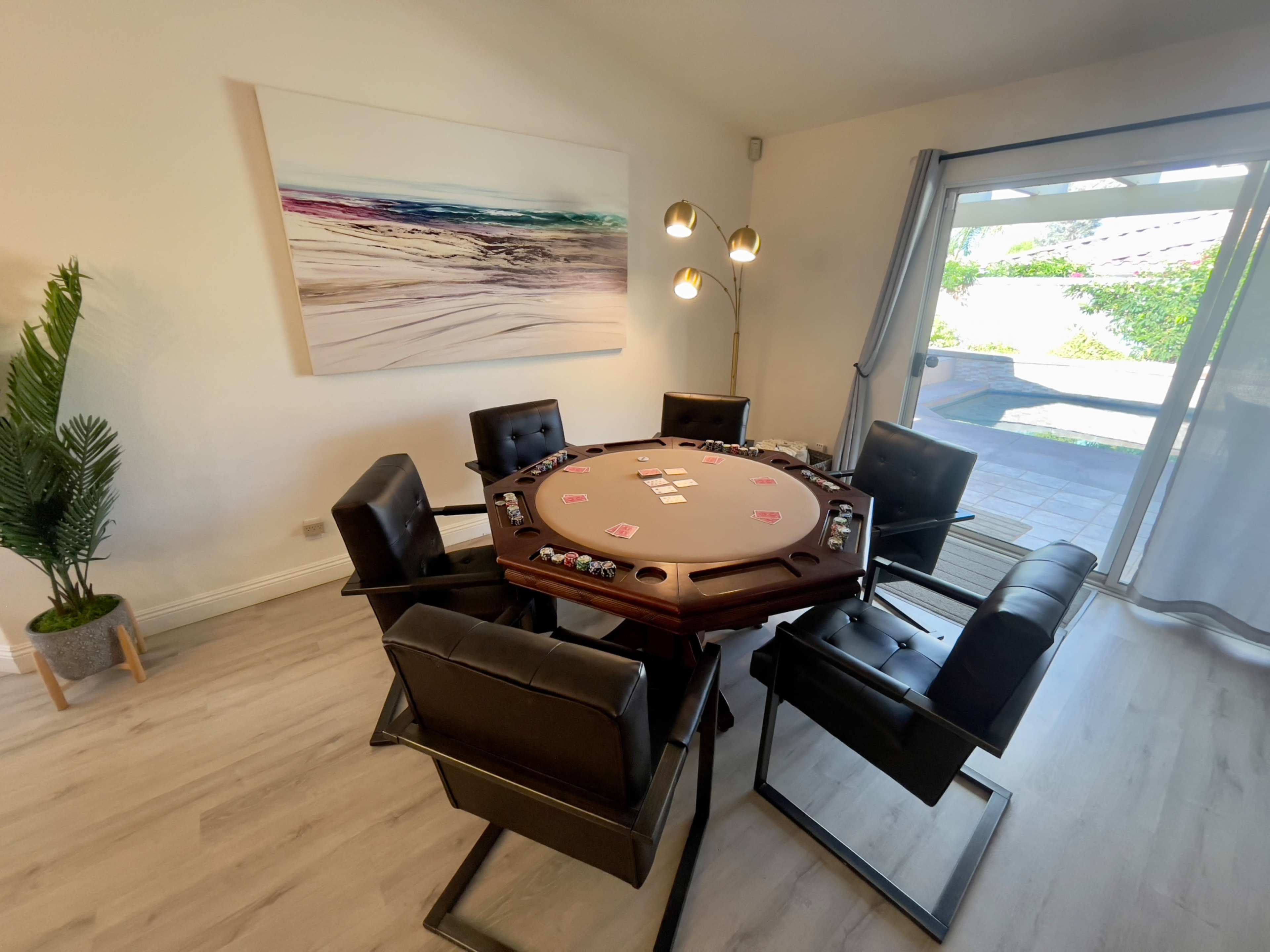 A round poker table is set with playing cards and chips, surrounded by six black chairs in a well-lit room with a large window overlooking a patio.