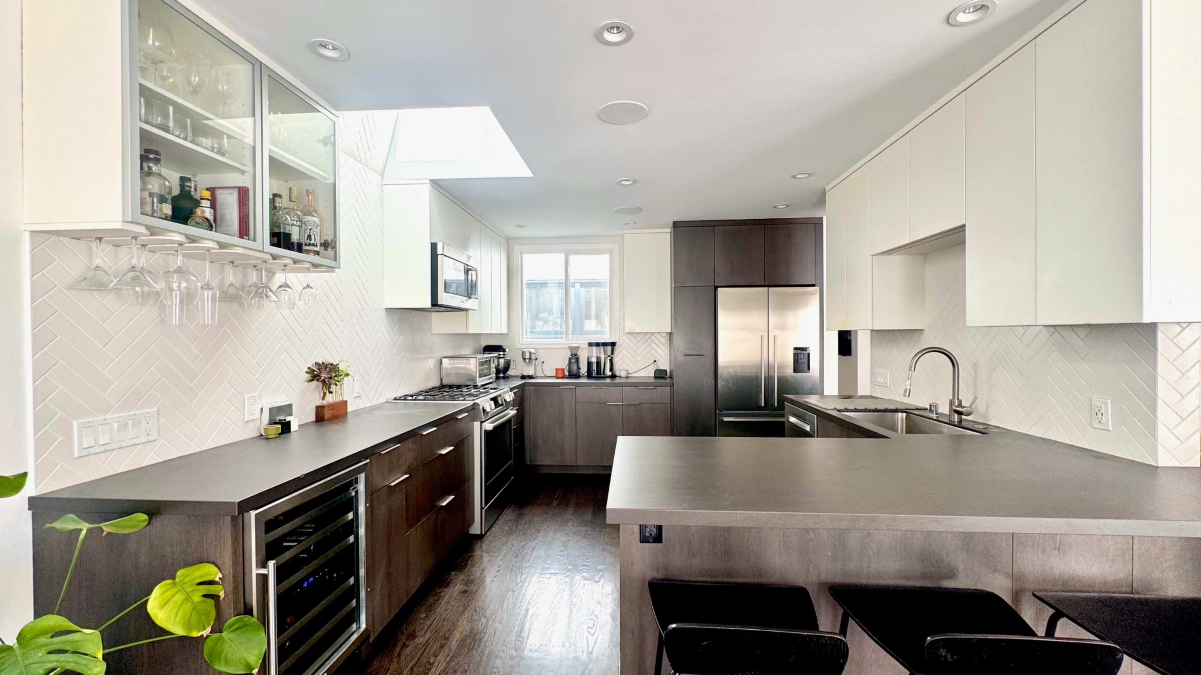 The image shows a modern kitchen with a combination of dark and light cabinetry, a central island, and large windows allowing natural light.
