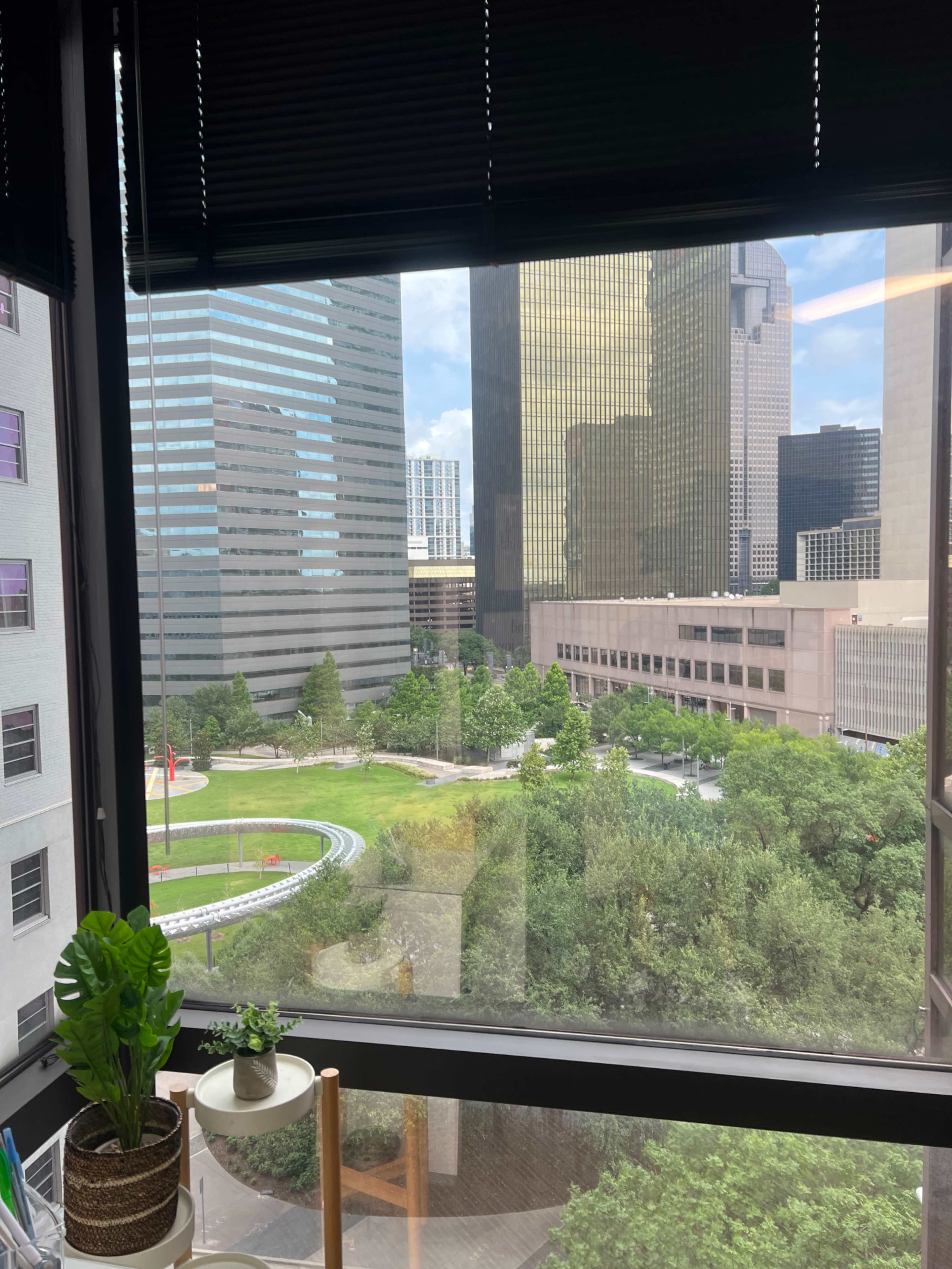 The image shows a view from an office window overlooking a green park area surrounded by tall commercial buildings in an urban setting.
