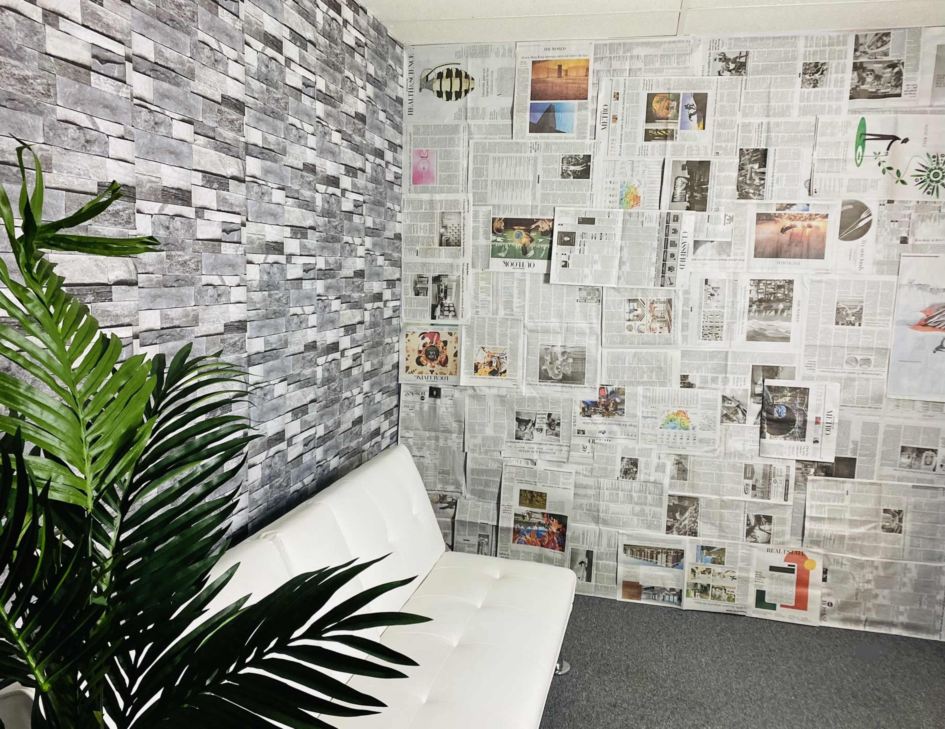 A room with one wall covered in gray stone wallpaper and the opposite wall decorated with newspaper clippings, featuring a white sofa and a green plant in the corner.