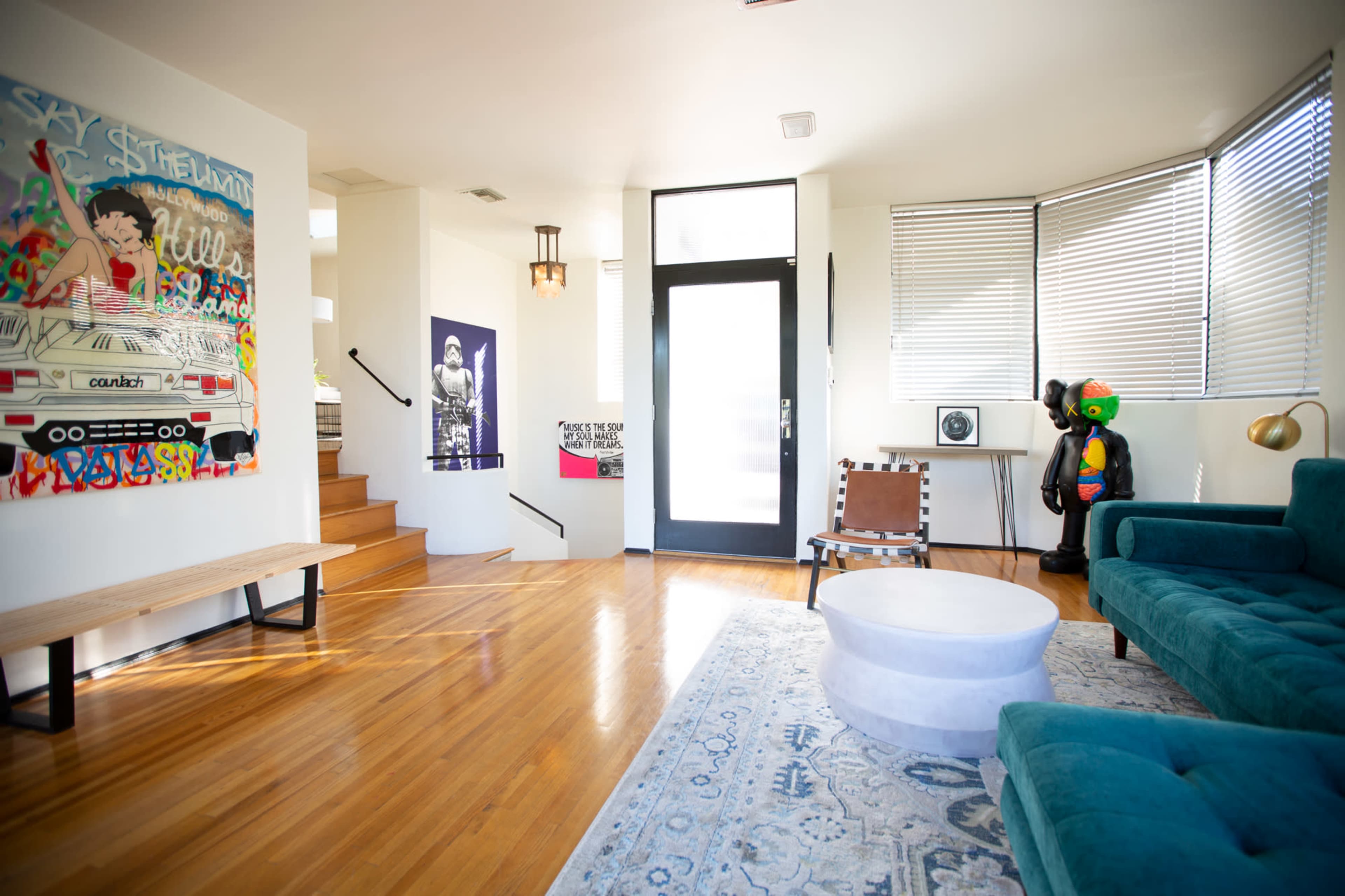 A modern living room features a teal sofa, a circular coffee table, artwork on the walls, and a staircase leading to an upper level.
