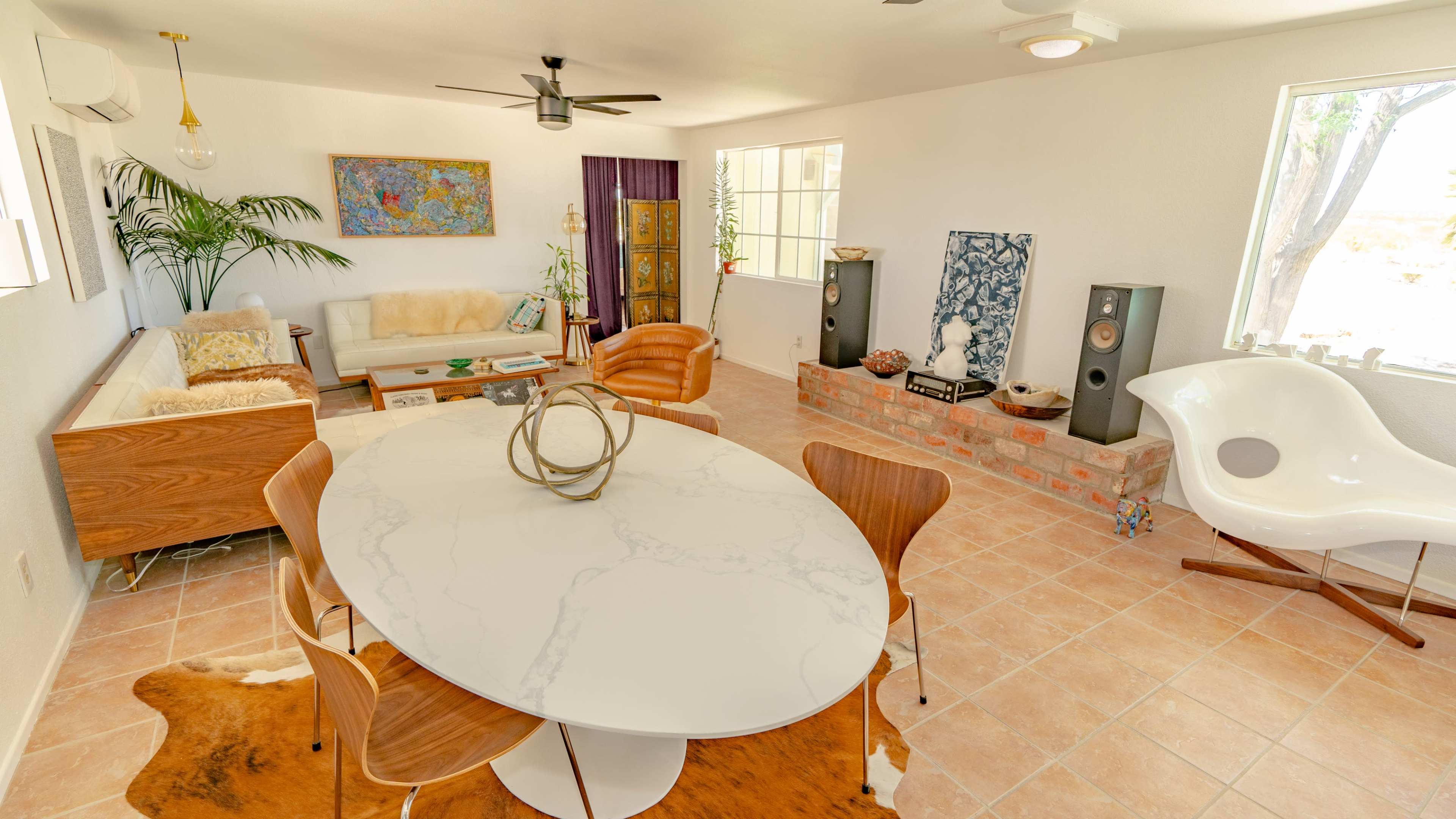 A brightly lit living space featuring a circular marble dining table, a wooden sofa, and modern chairs, with large windows and decorative elements such as plants and art.