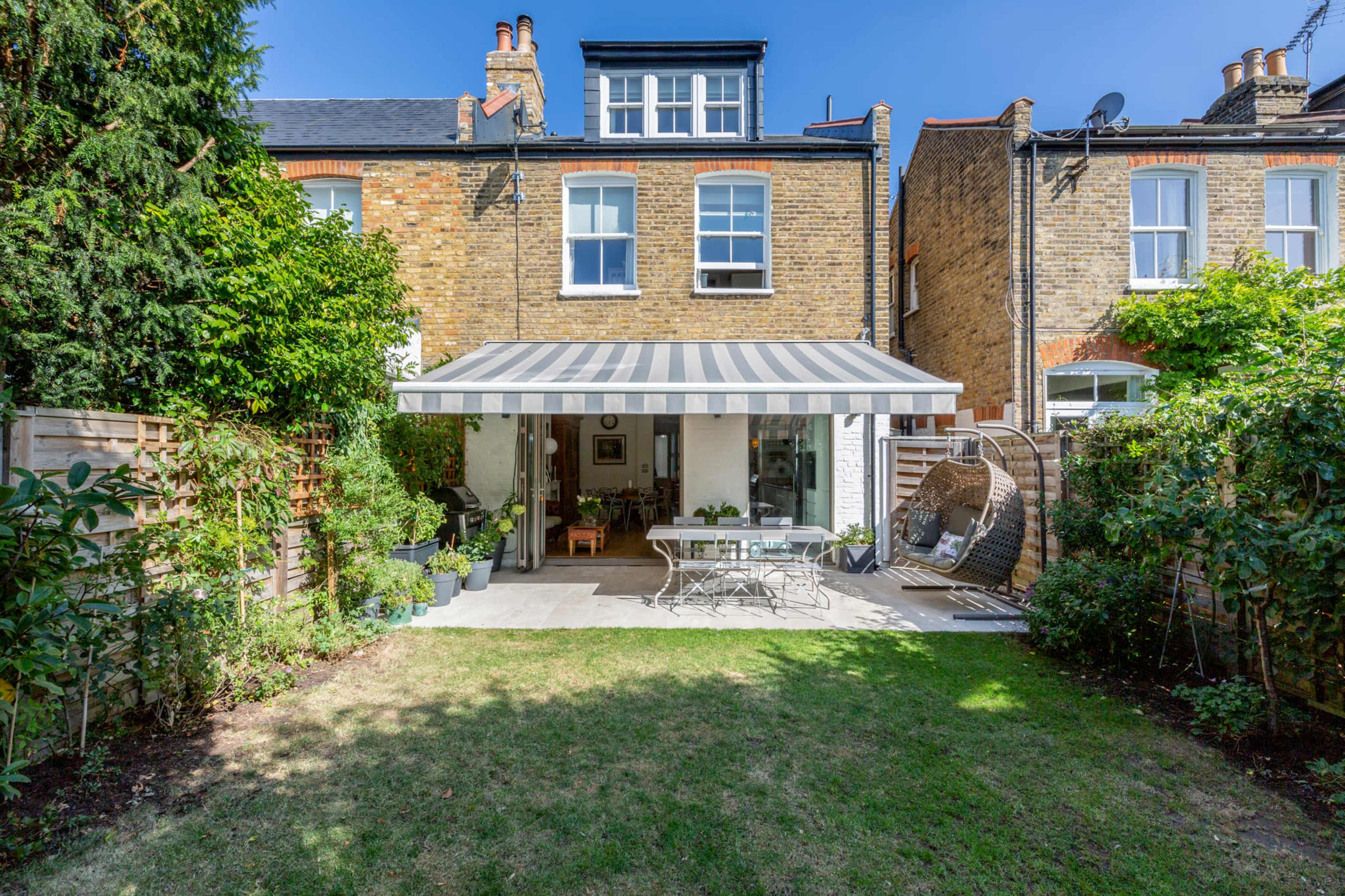 The image shows a backyard with a patio featuring a striped awning, a table and chairs, and well-maintained greenery surrounding a brick house.