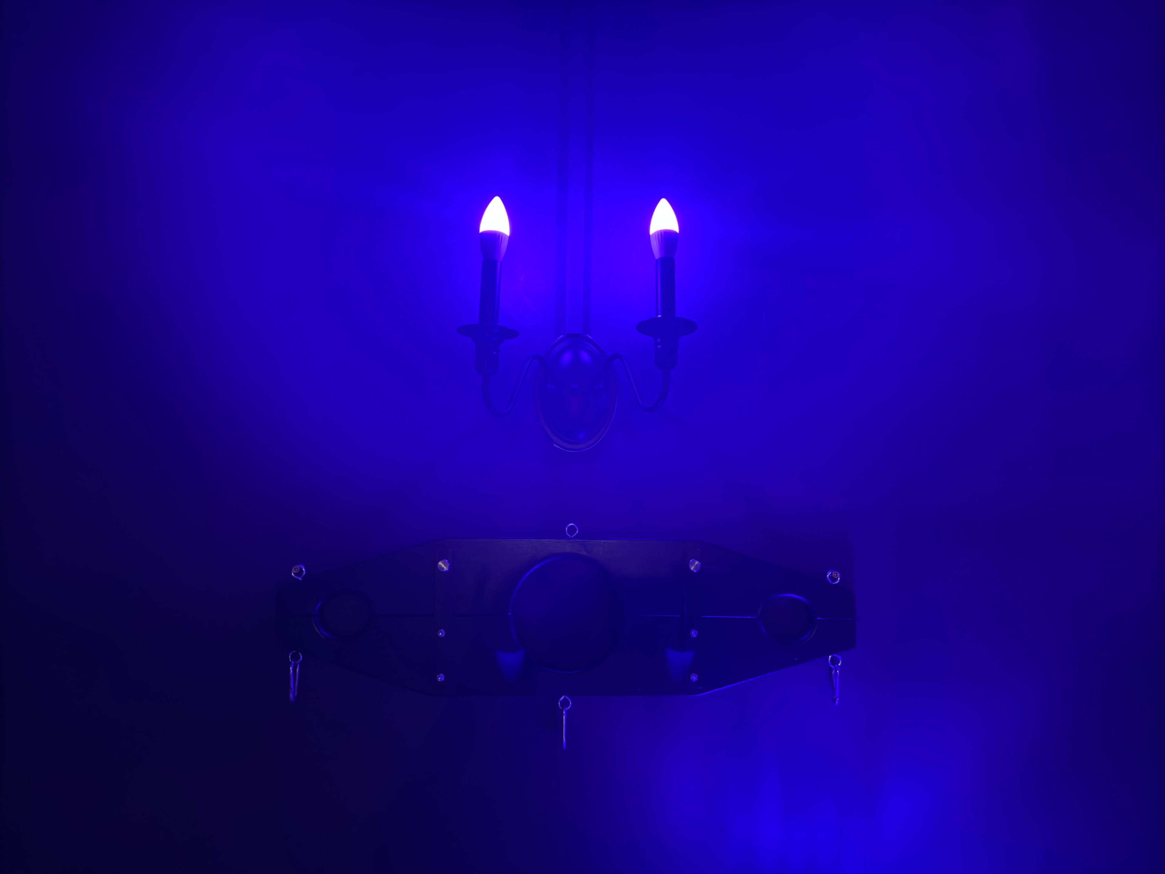 The image features a wall with a dark background, adorned with a black apparatus and two flickering candle-like lights emitting a bluish glow.