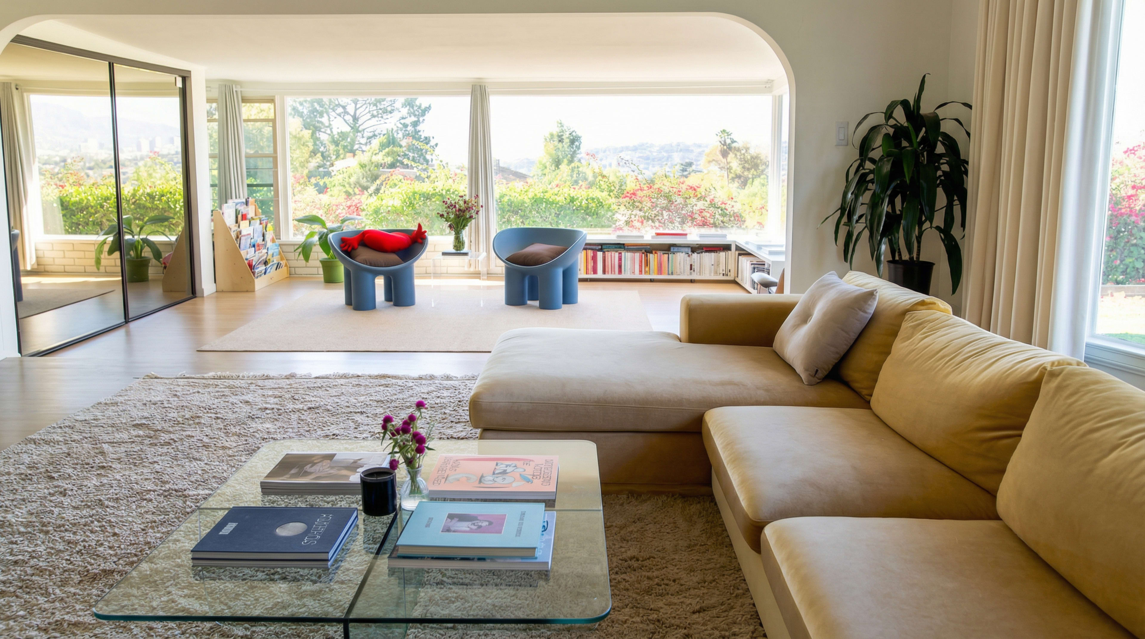 The image shows a modern living room with a beige sofa, a glass coffee table, and two colorful chairs, surrounded by large windows that offer a view of a garden.