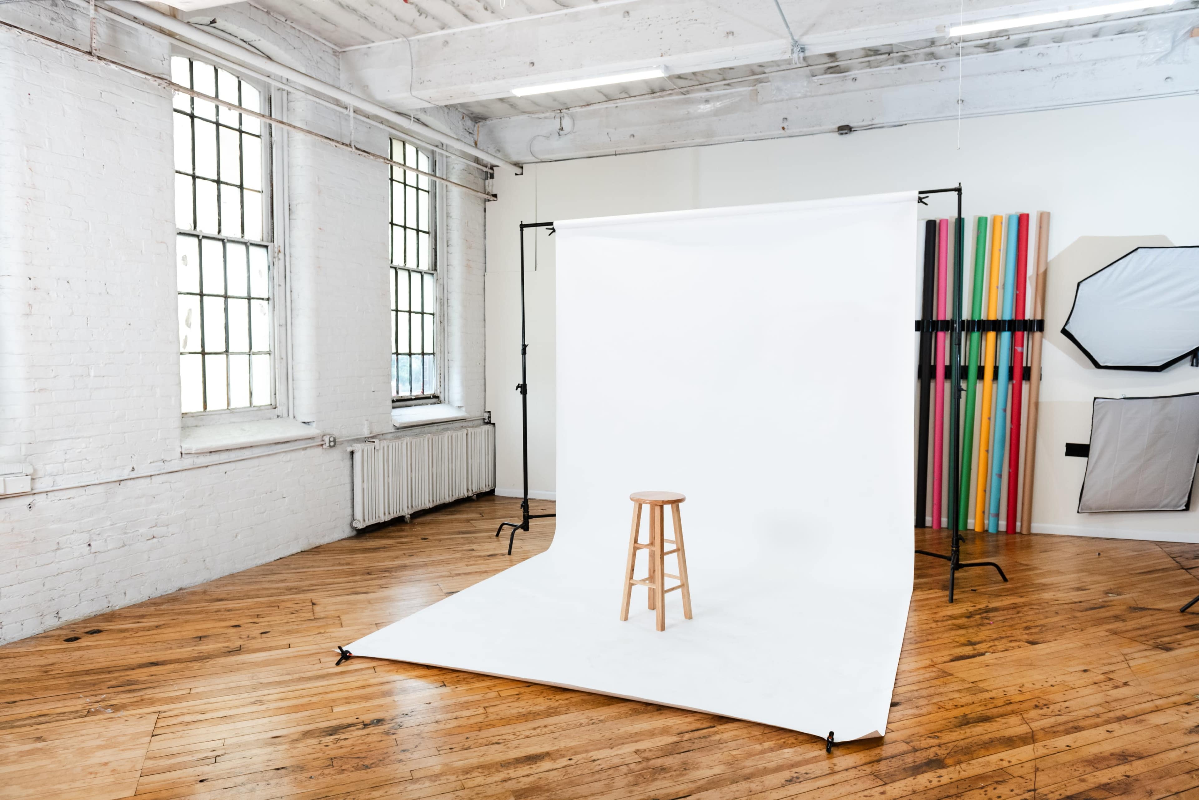 A photography studio with a wooden stool positioned on a white backdrop, surrounded by large windows and colorful props.