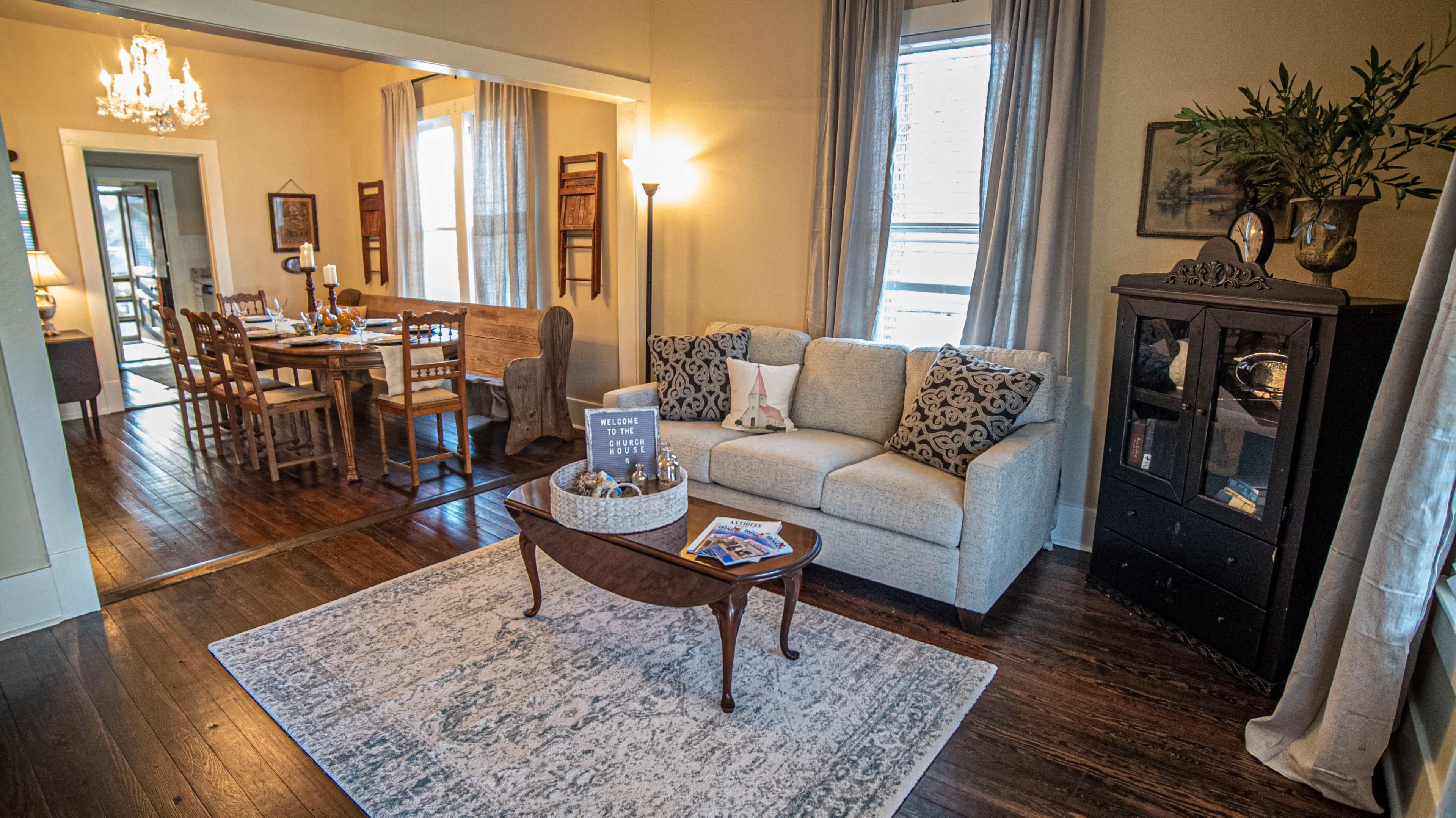 A cozy living area with a light gray sofa, a decorative coffee table, and a dining area visible in the background with a wooden table set for a meal.