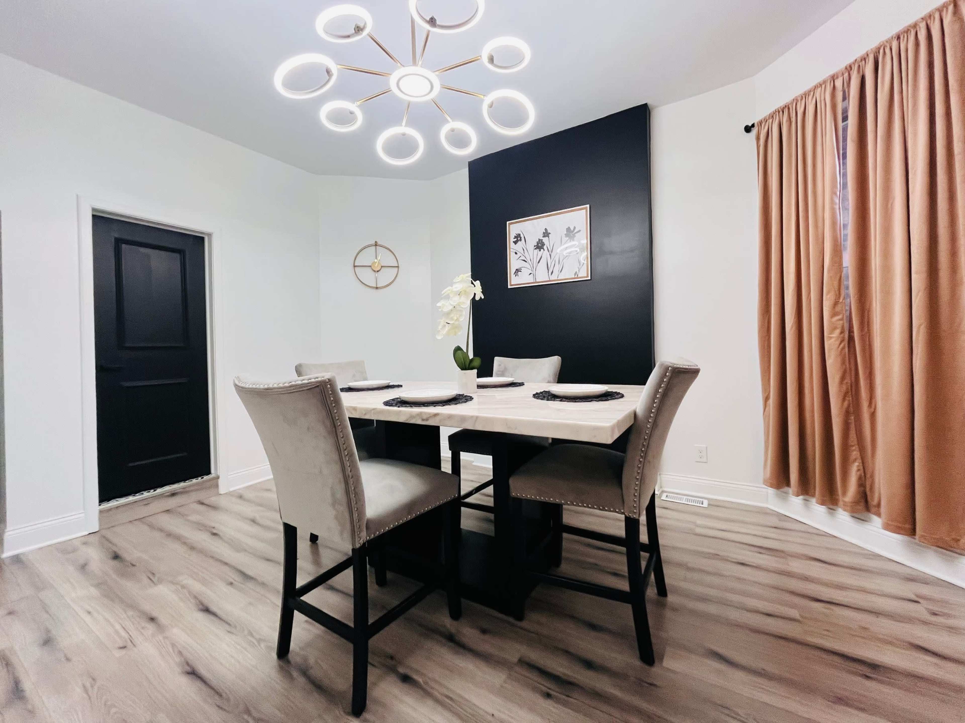 A dining area features a table surrounded by four chairs, with a circular chandelier above and a black accent wall behind, complemented by a window draped with a curtain.