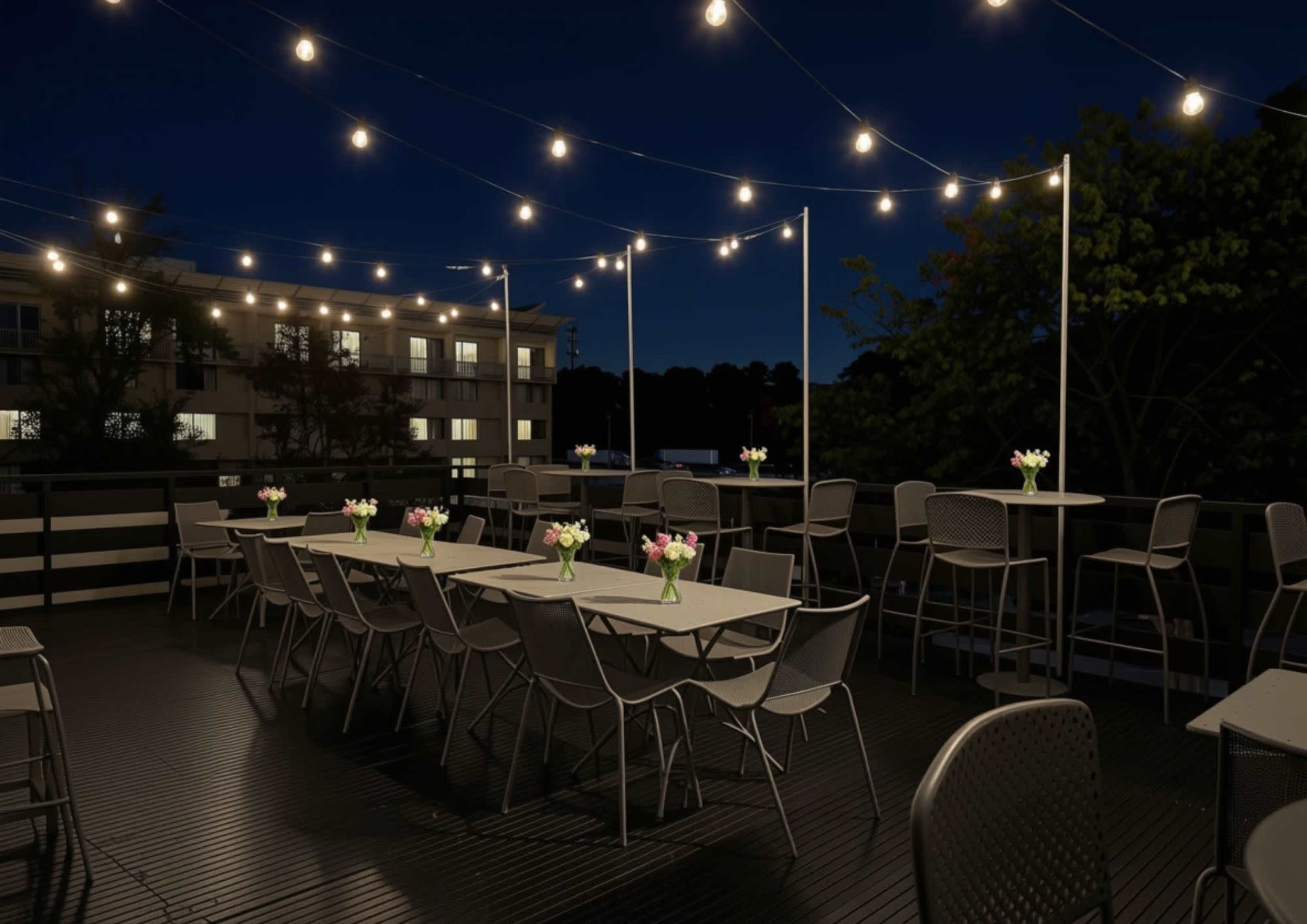 The image shows a rooftop dining area at night, featuring tables with flower centerpieces and string lights illuminating the space.