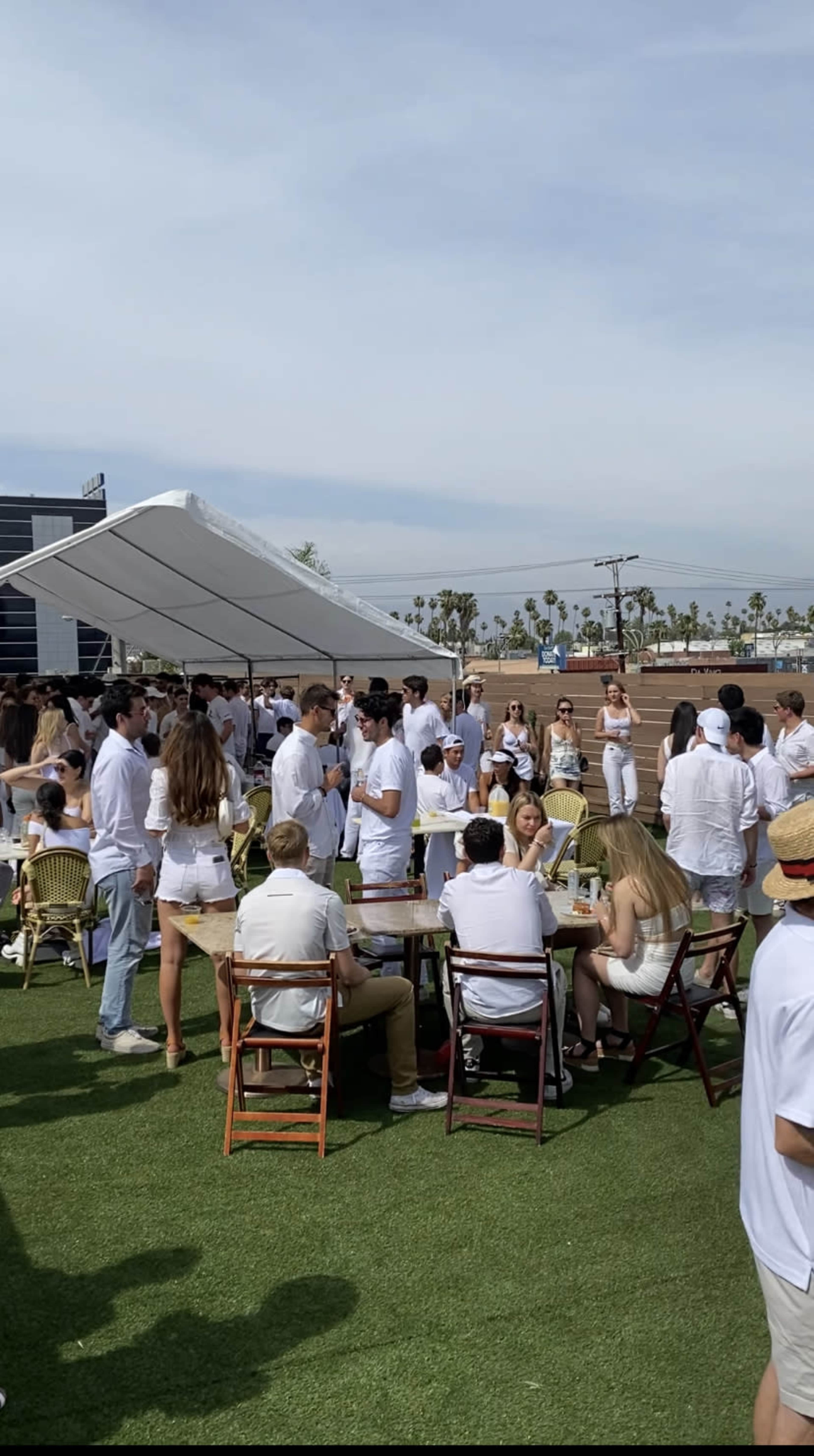 A large group of people dressed in white gathers outdoors on a green lawn with tables and a tent.