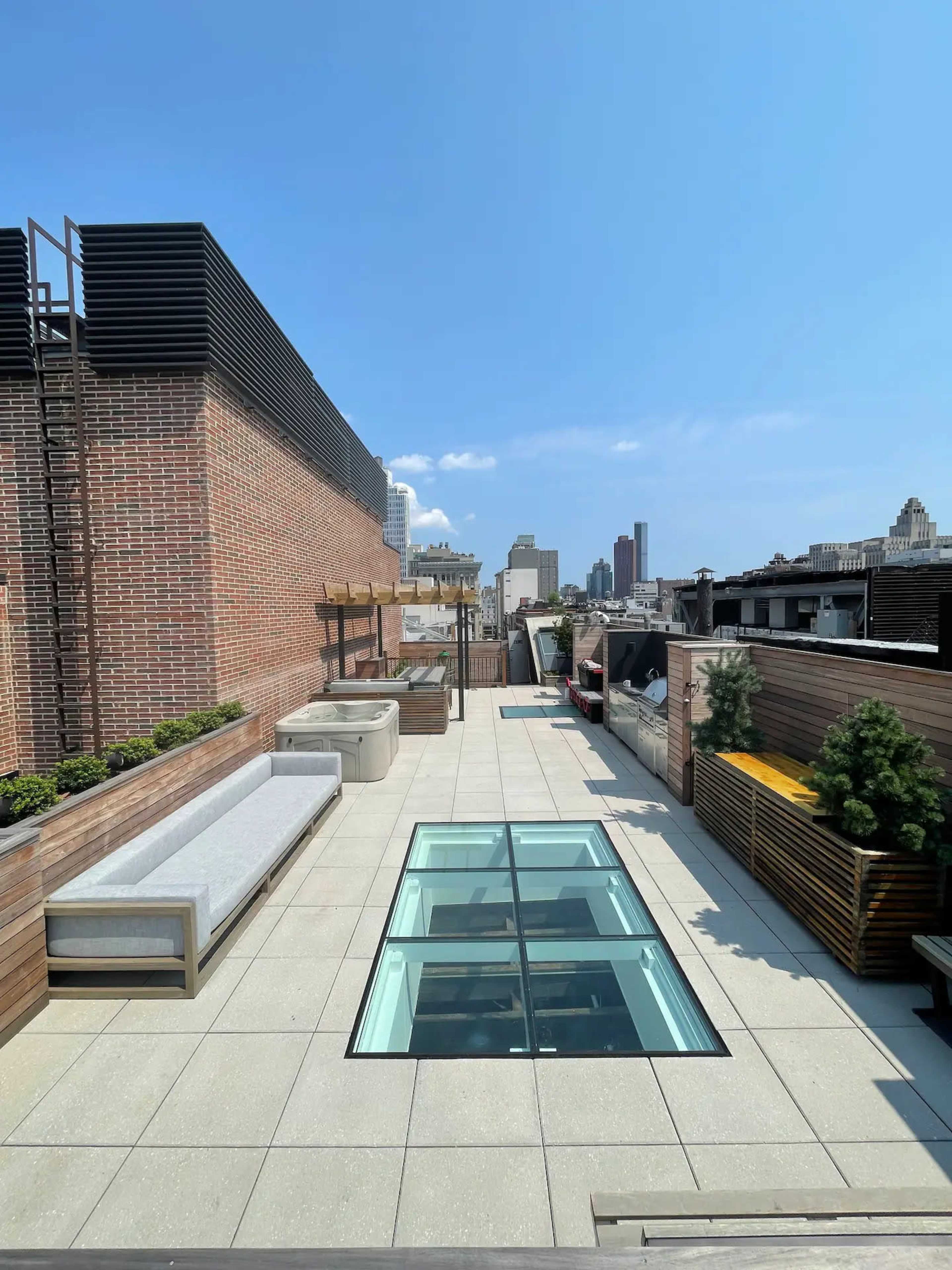 A rooftop terrace featuring glass panels in the center, surrounded by seating areas, planters, and a skyline view.