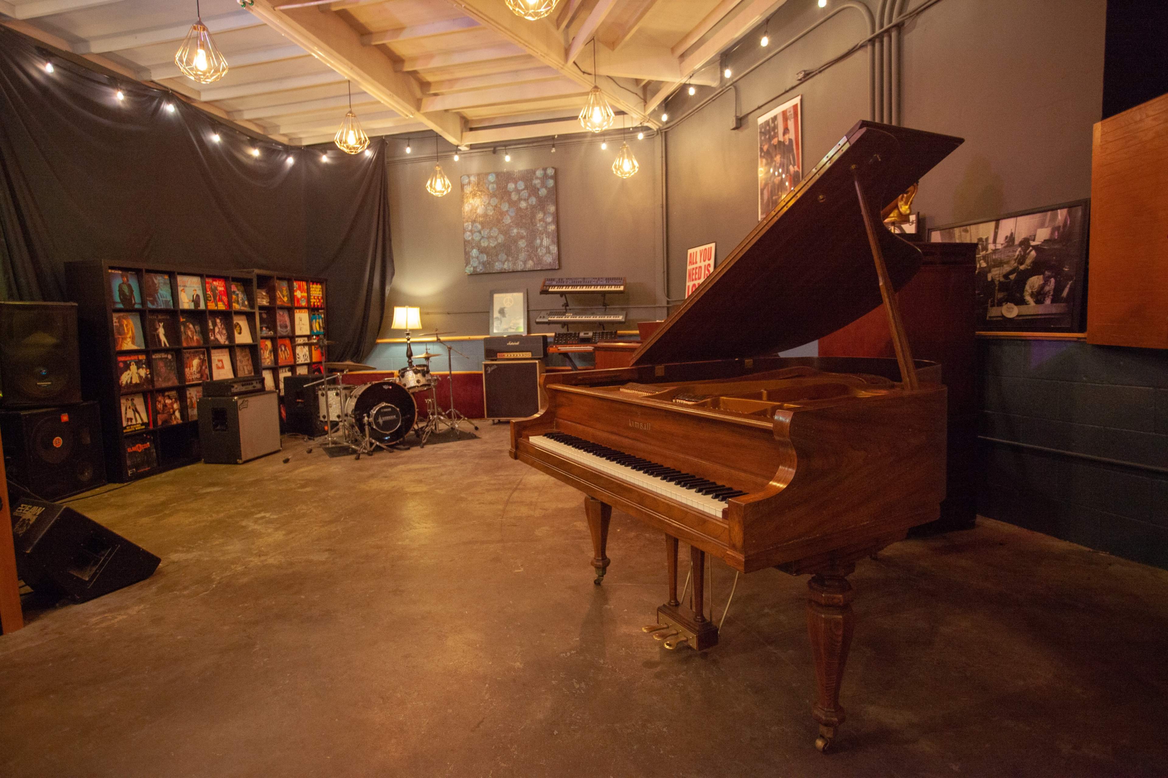 A music studio featuring a grand piano, a drum set, speakers, record shelves, and warm overhead lights.