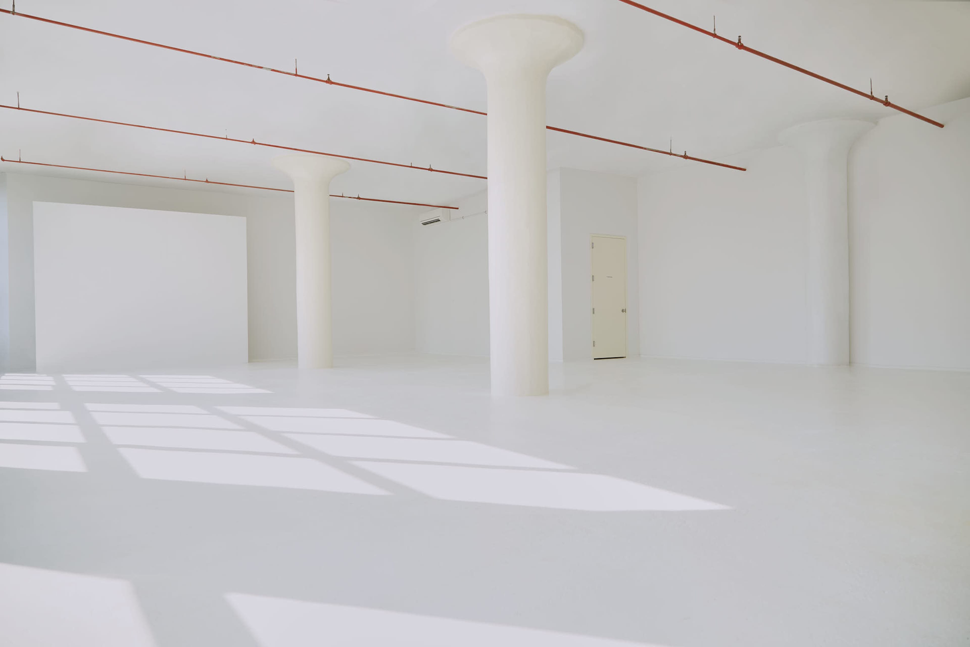 An empty white room with concrete pillars and large windows casting shadows on the floor.