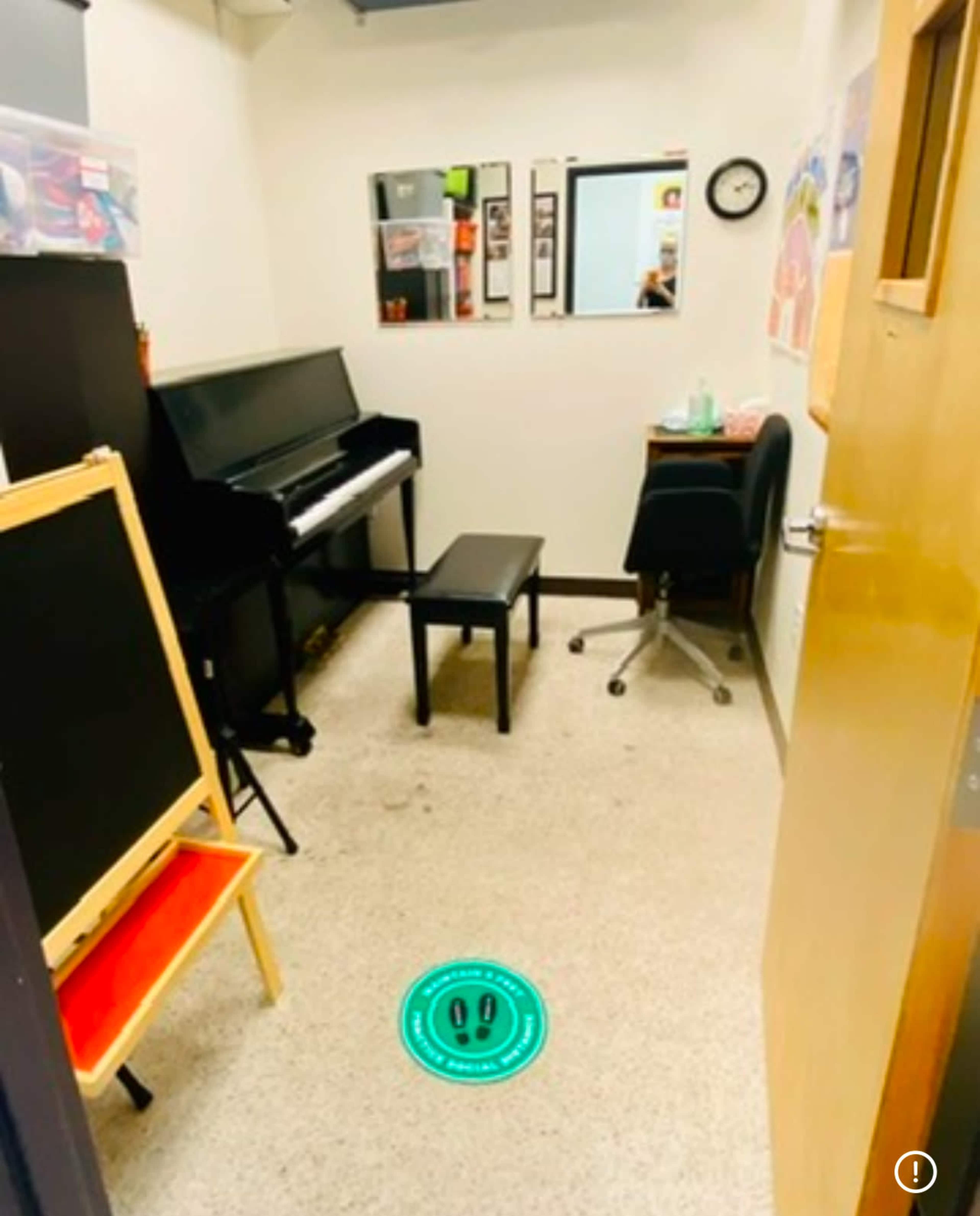 The image shows a small music practice room with a black piano, a bench, a chair, a chalkboard, and a clock on the wall.