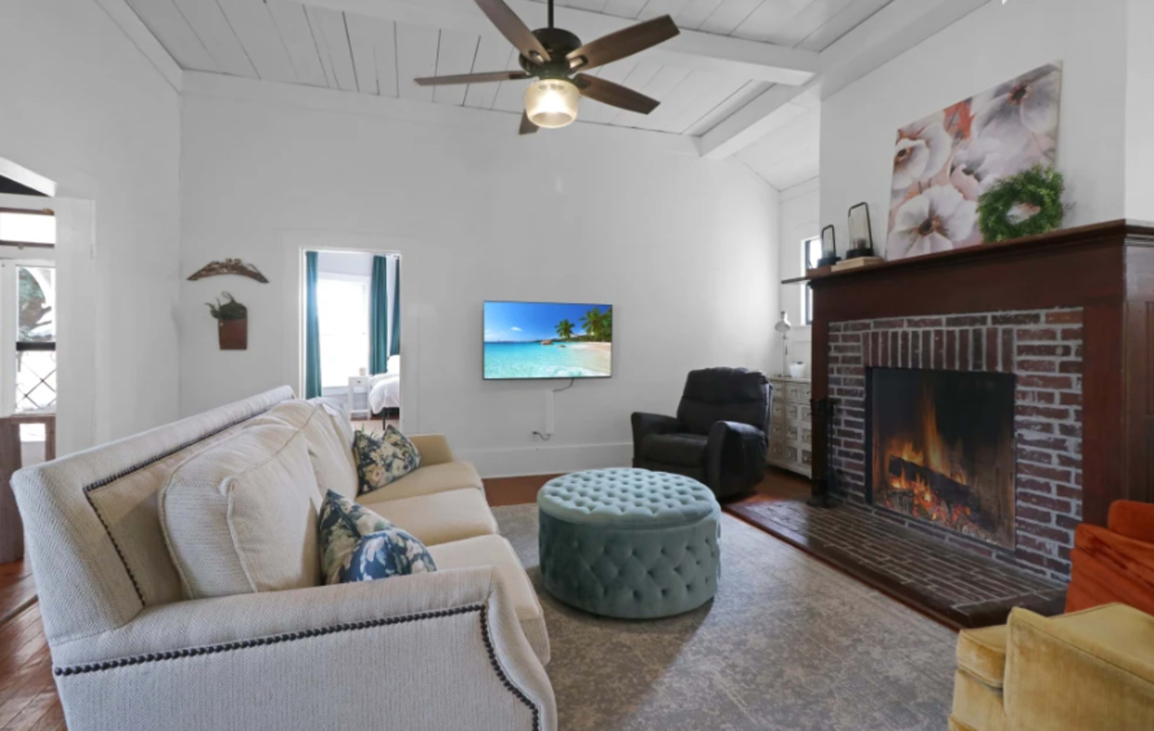 The living room features a white sofa, a teal ottoman, a brick fireplace with an accompanying television mounted on the wall, and a ceiling fan.
