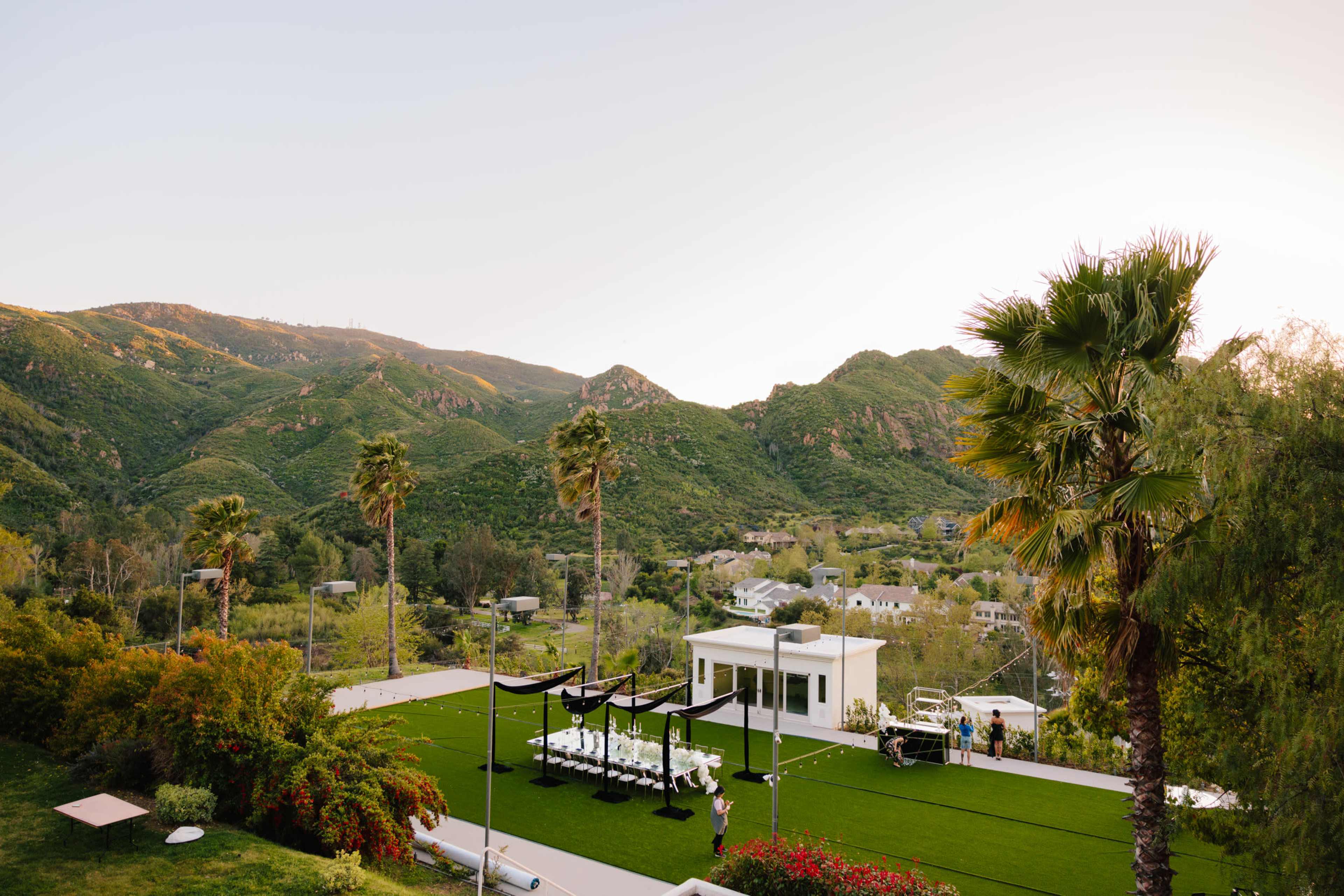 A modern outdoor event space is set up on a grassy area, surrounded by palm trees and hills in the background.