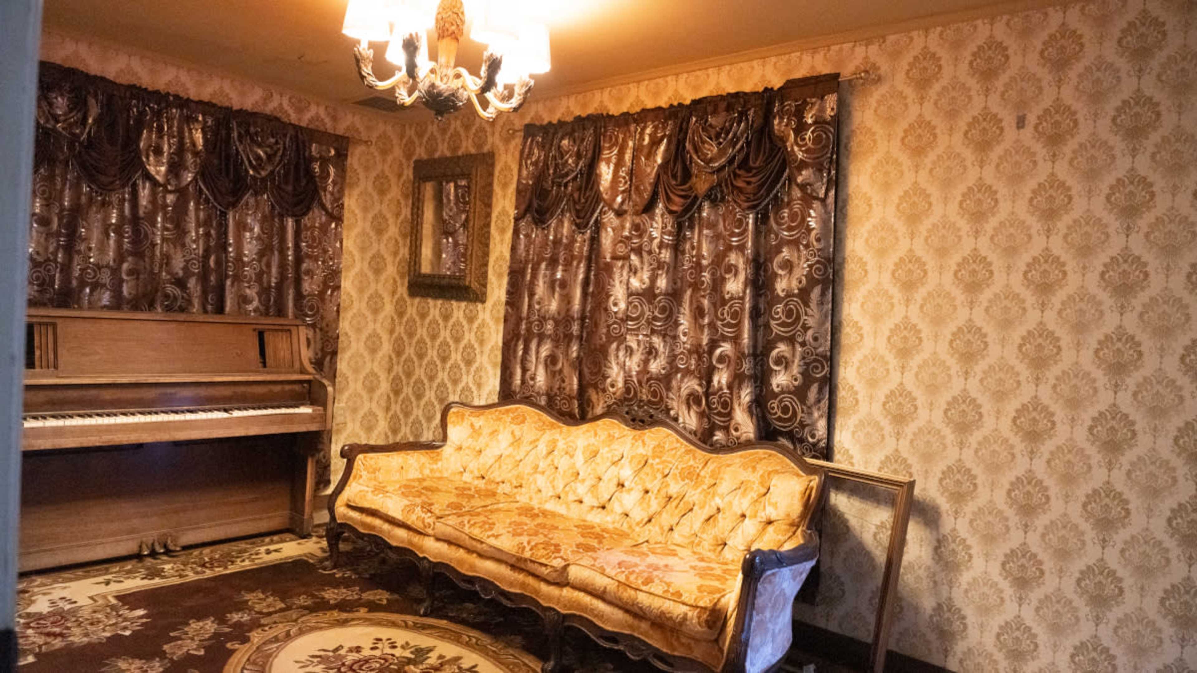 The room features a vintage piano, a decorative sofa with ornate upholstery, and patterned curtains against a floral wallpaper backdrop.