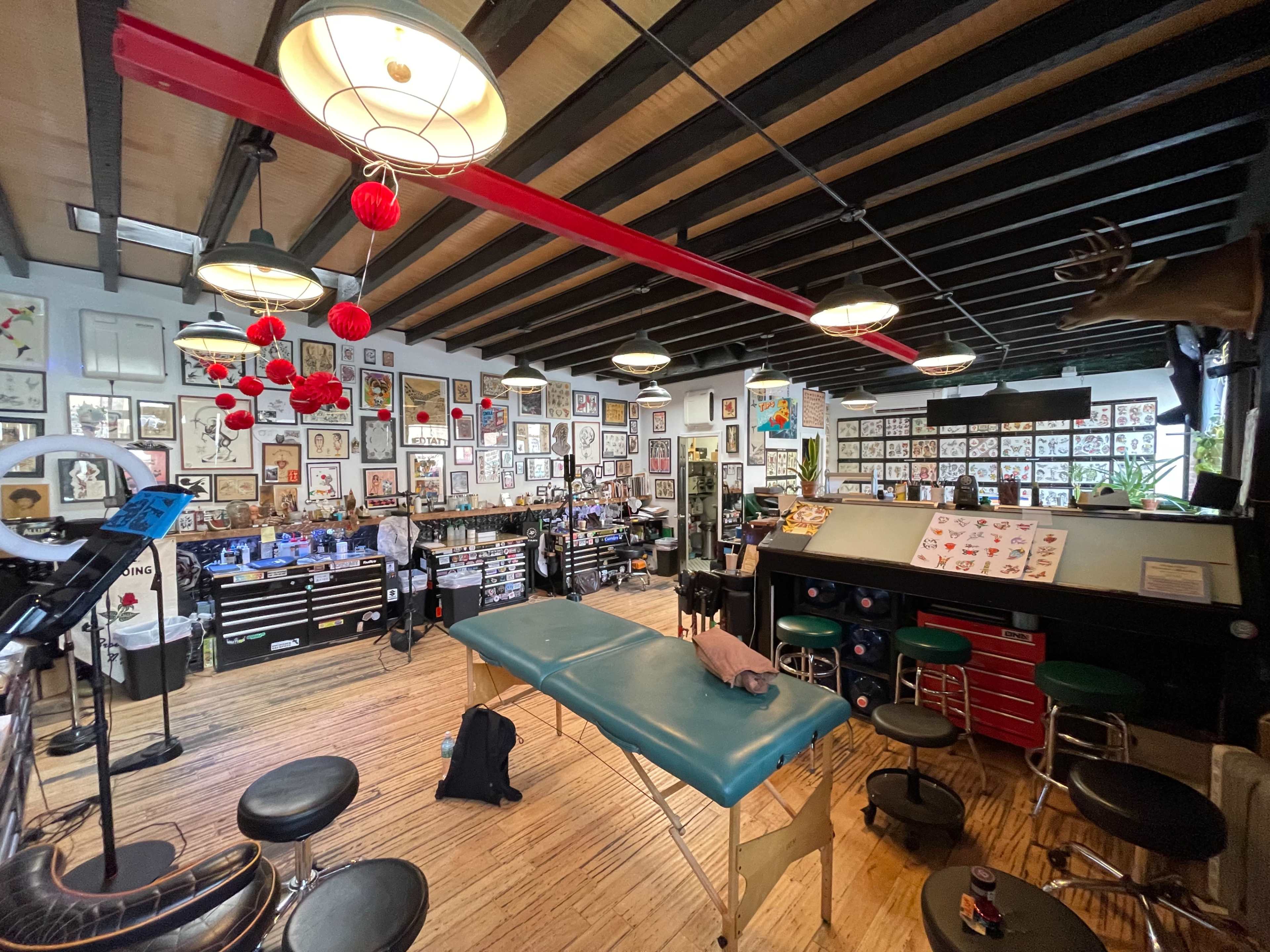 A tattoo studio features a variety of tattoo designs displayed on the walls, tattooing equipment organized on counters, and a green tattoo table in the center.