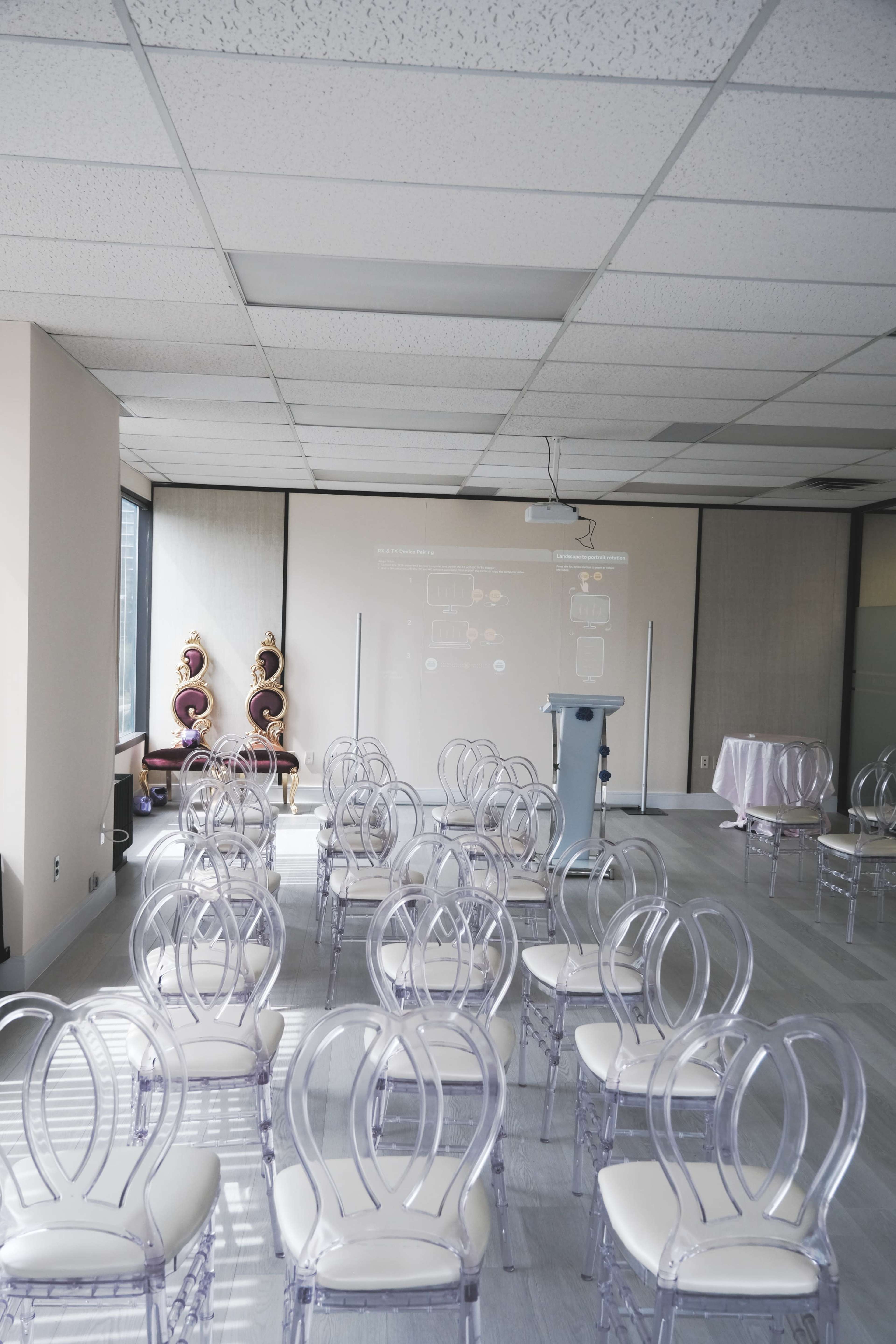 A spacious room is set up with rows of clear acrylic chairs facing a projector screen and a podium, with a decorative setup in the corner.