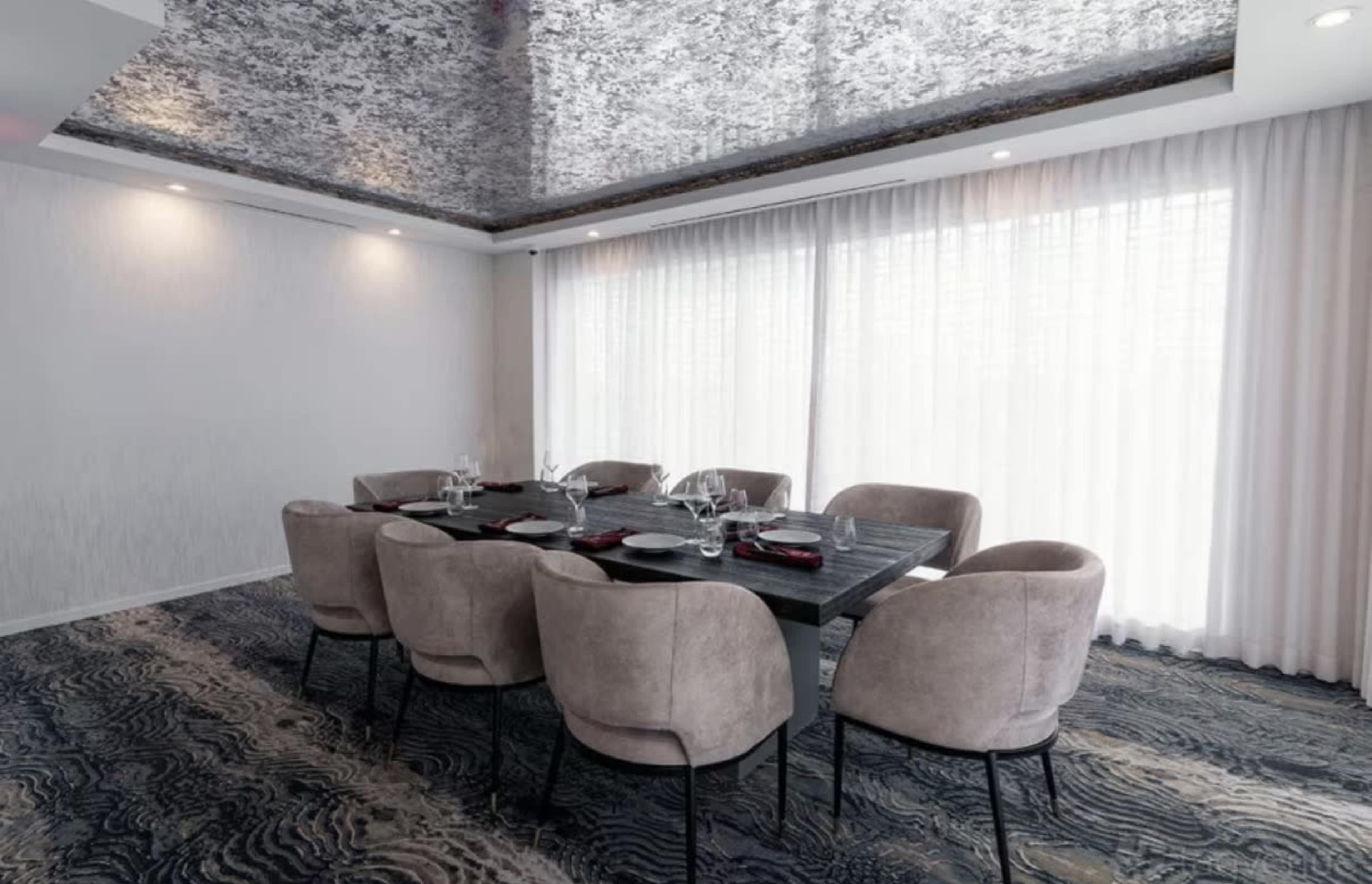 The image shows a modern dining room with a large rectangular table surrounded by eight upholstered chairs, set with tableware, and illuminated by natural light through sheer curtains.