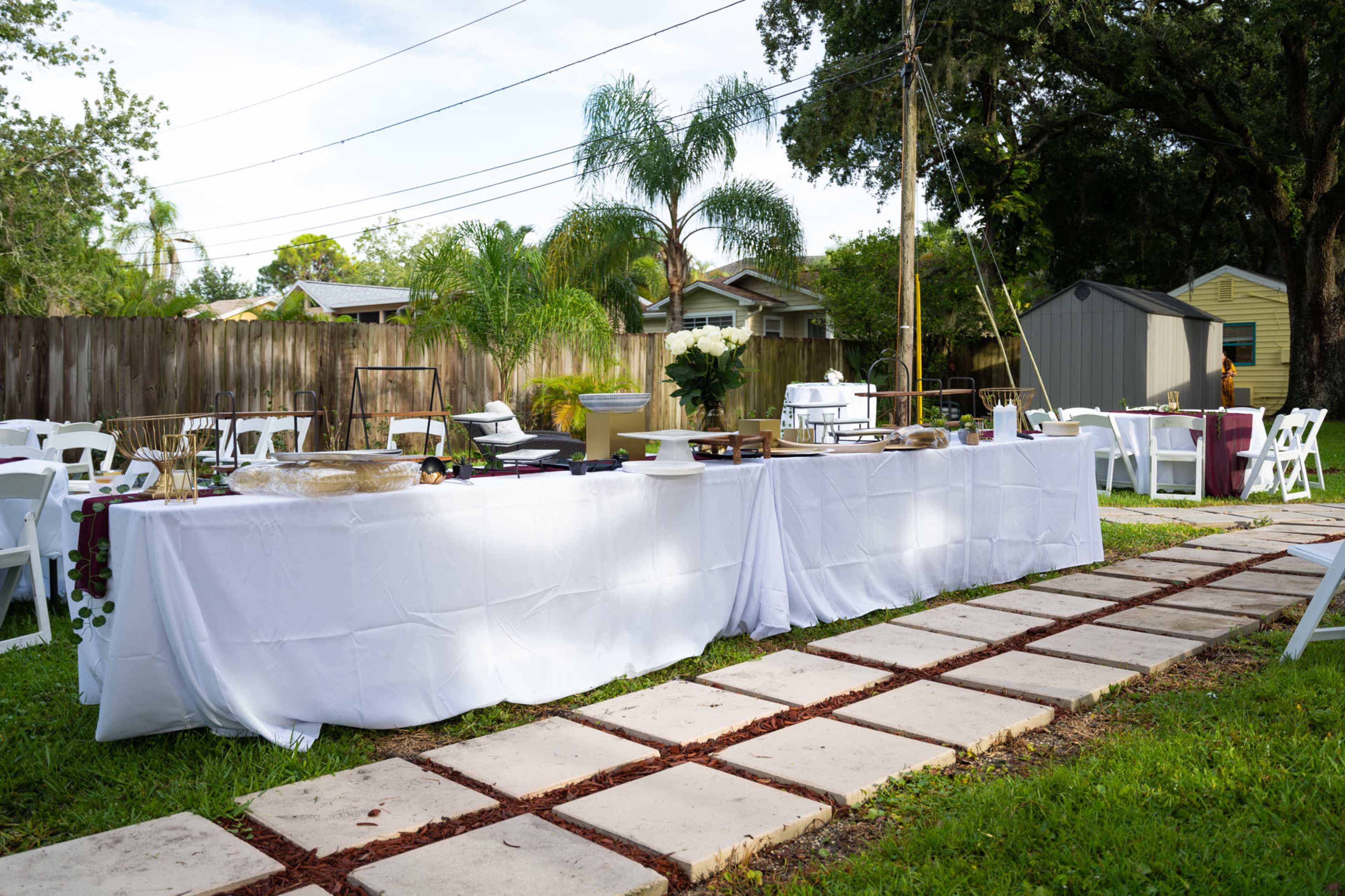 A white tablecloth-covered table is set up outdoors, surrounded by chairs and a stone pathway in a backyard with trees and a shed in the background.