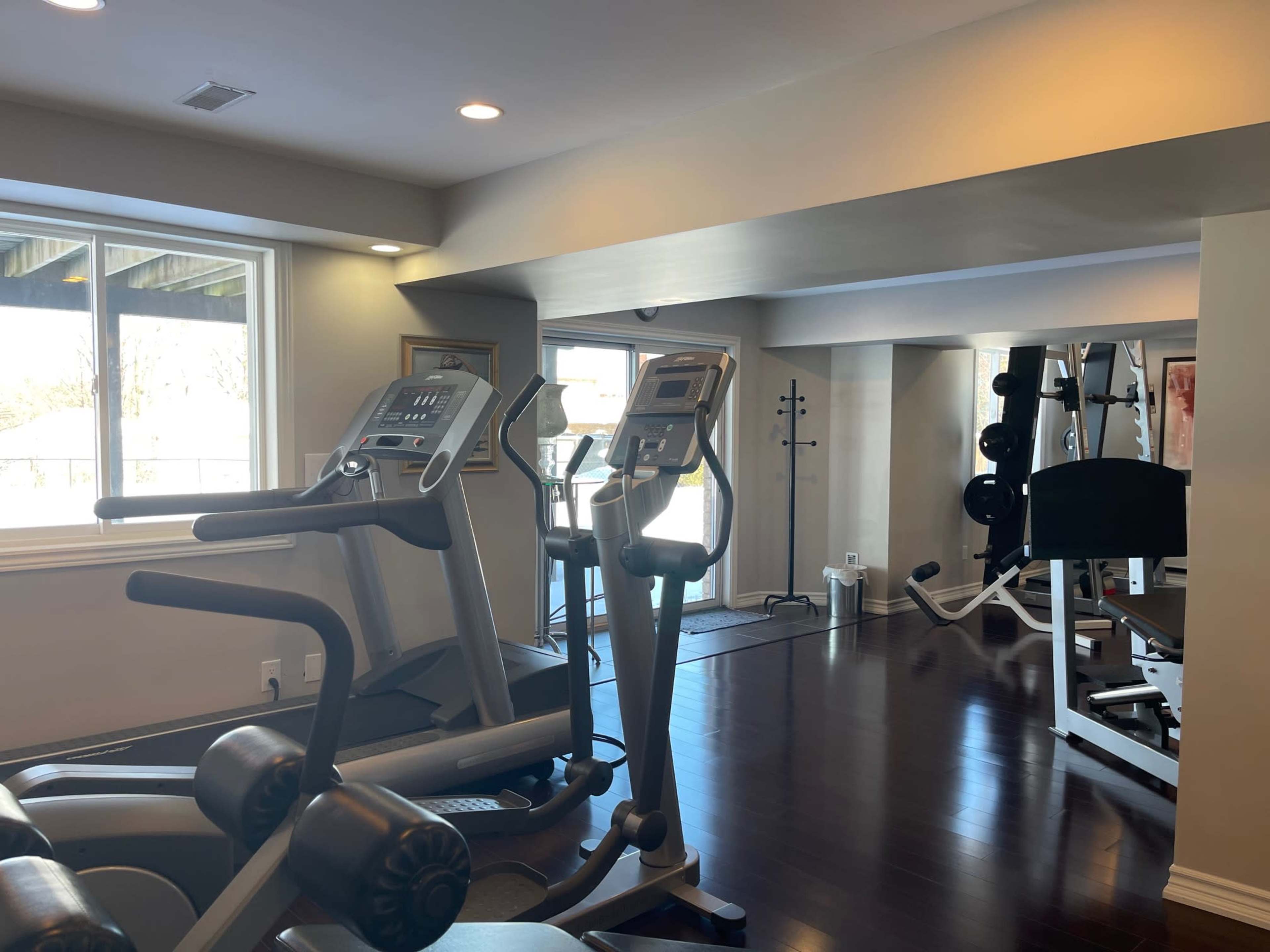 The image shows a fitness room equipped with exercise machines, including treadmills and weightlifting equipment, with large windows letting in natural light.