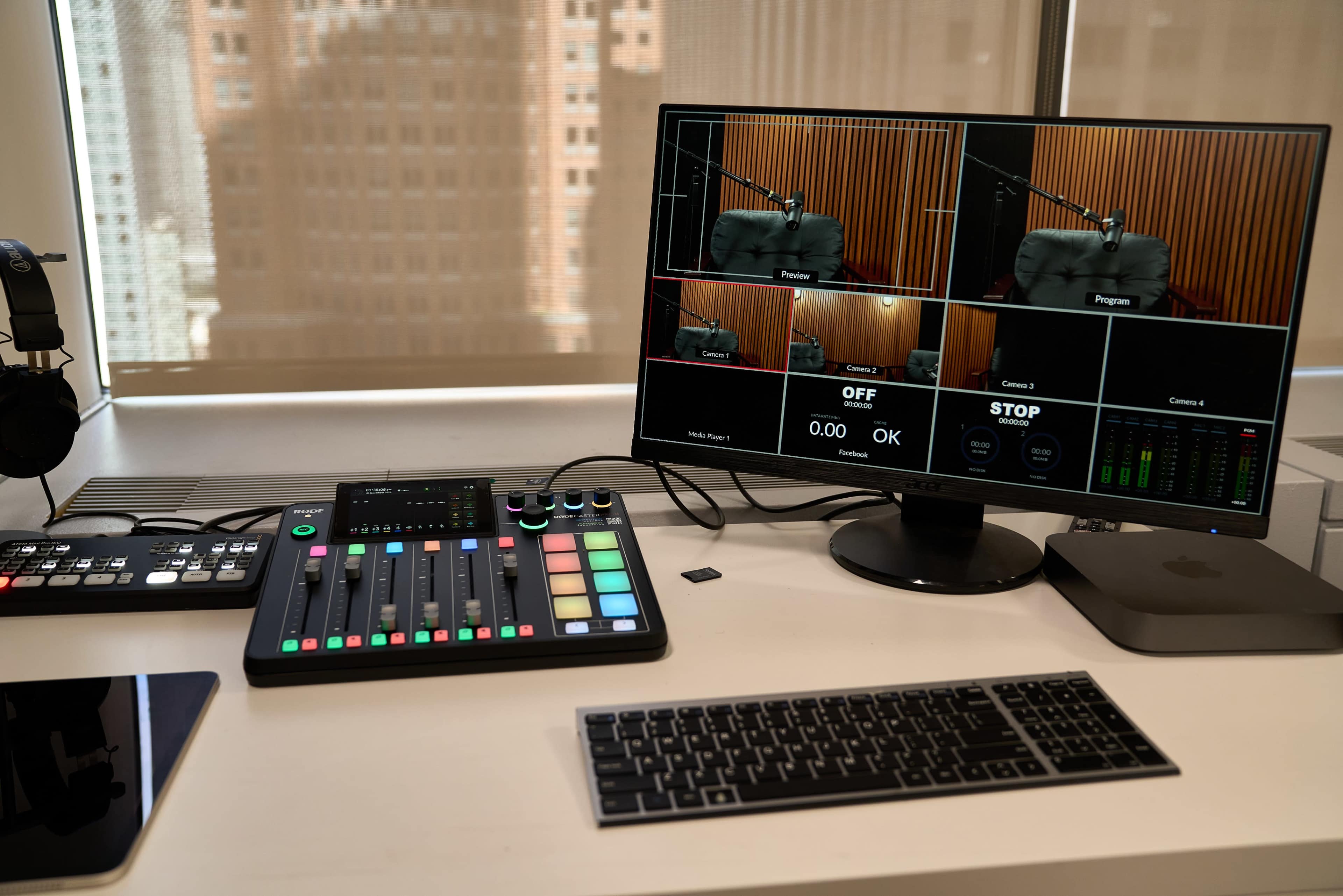 A recording studio setup features a control panel with colorful buttons, a computer monitor displaying multiple camera feeds, a keyboard, and headphones beside an iPad.