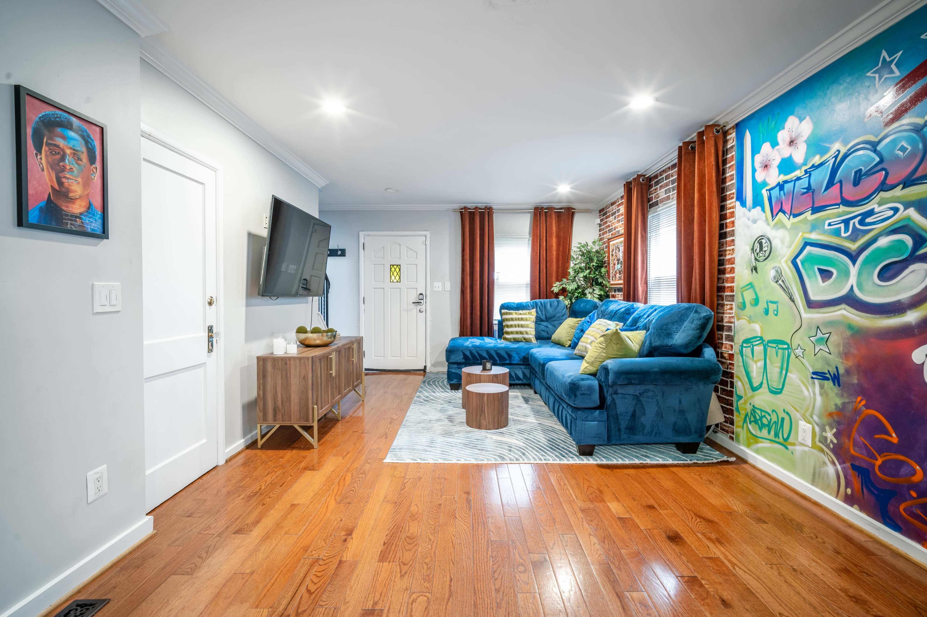 A modern living room featuring a blue sectional sofa, a wooden TV stand, and a colorful mural on a brick wall.