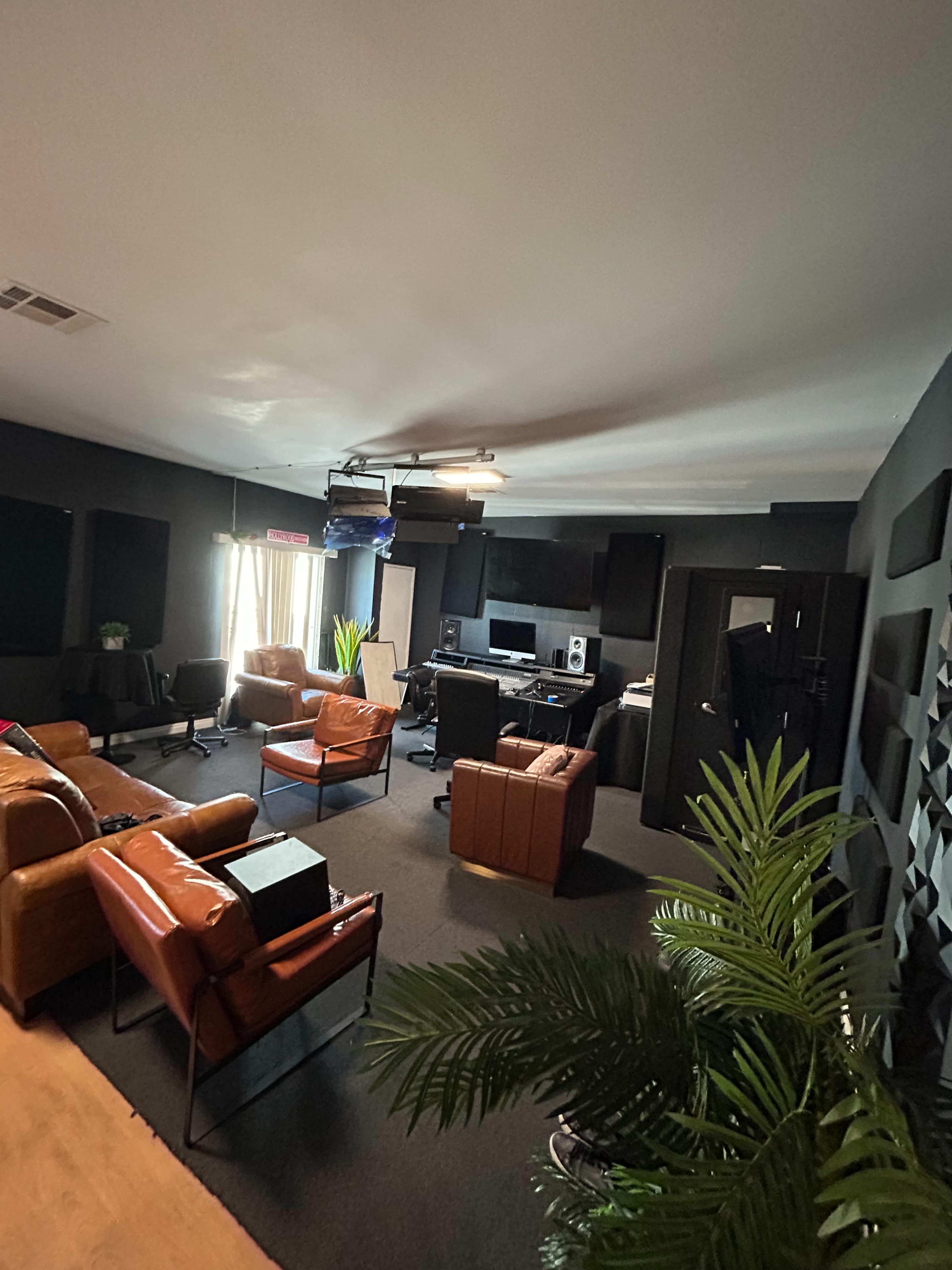 The image shows a modern recording studio with brown leather chairs, a computer setup, and large windows allowing natural light to enter.