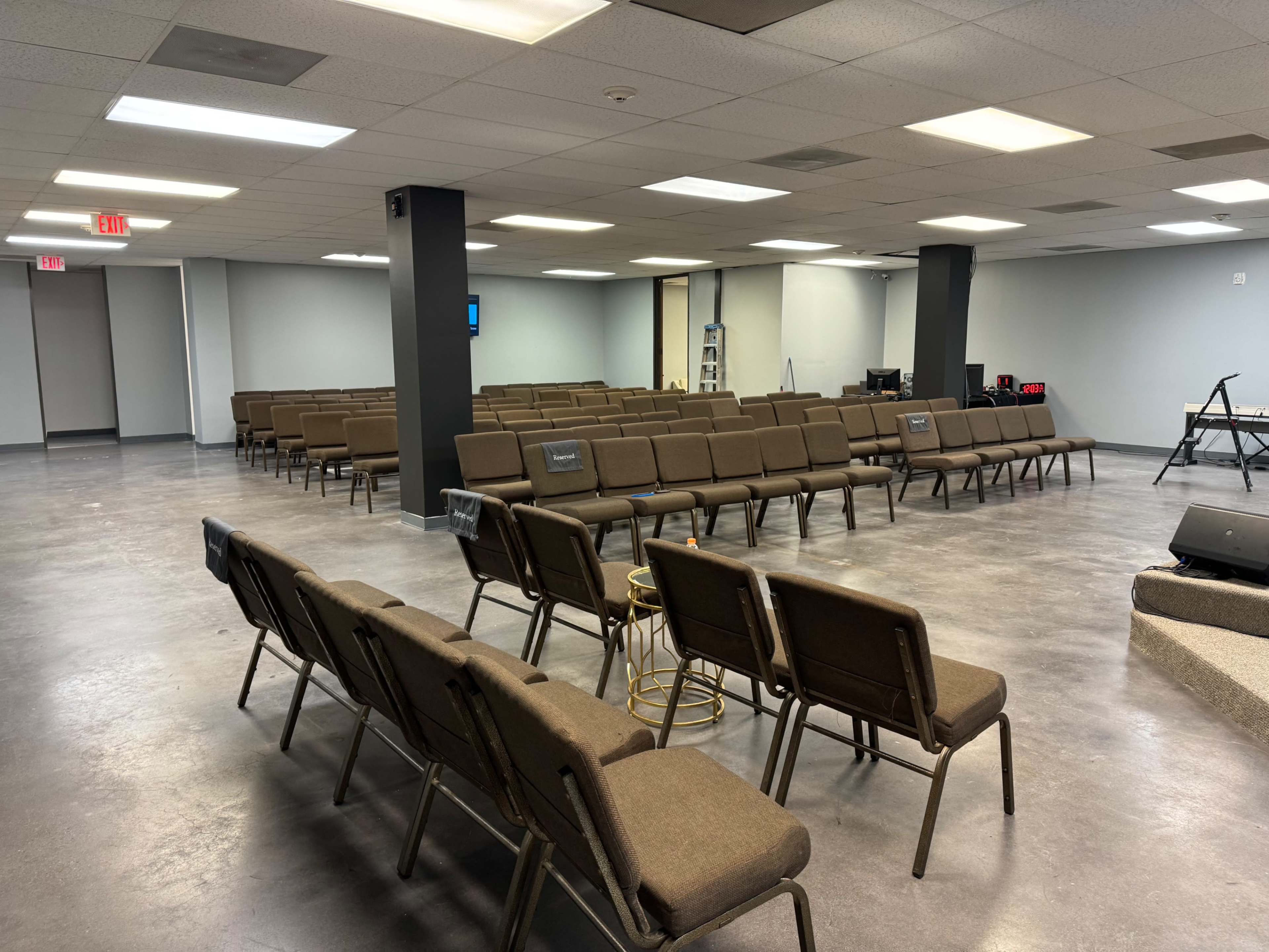 The image shows a large, empty room set up with rows of brown chairs facing a stage area.