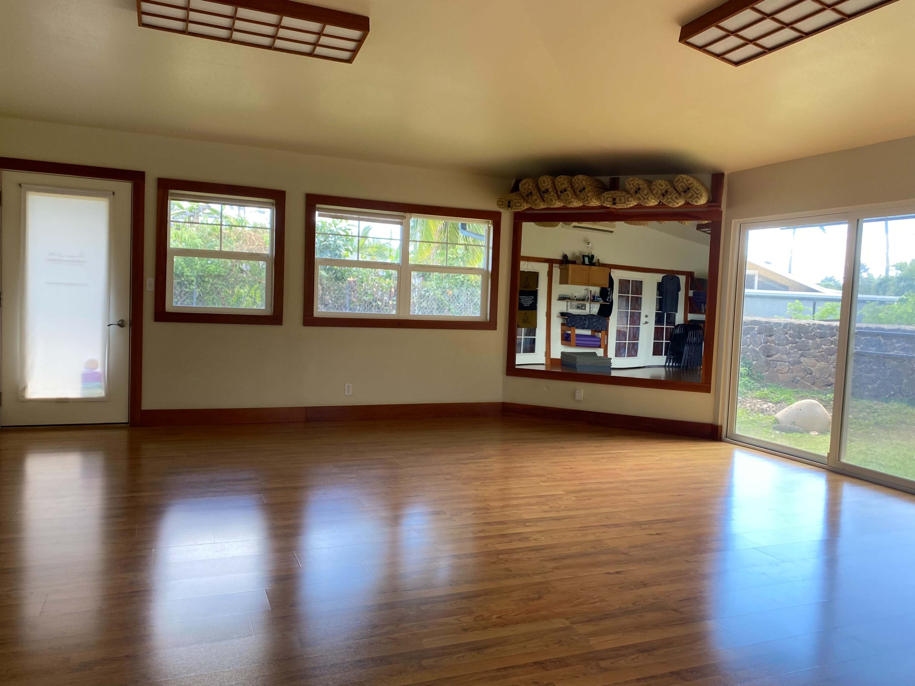 A spacious, well-lit room with wooden flooring, large windows on two walls, and a mirror reflecting the space.