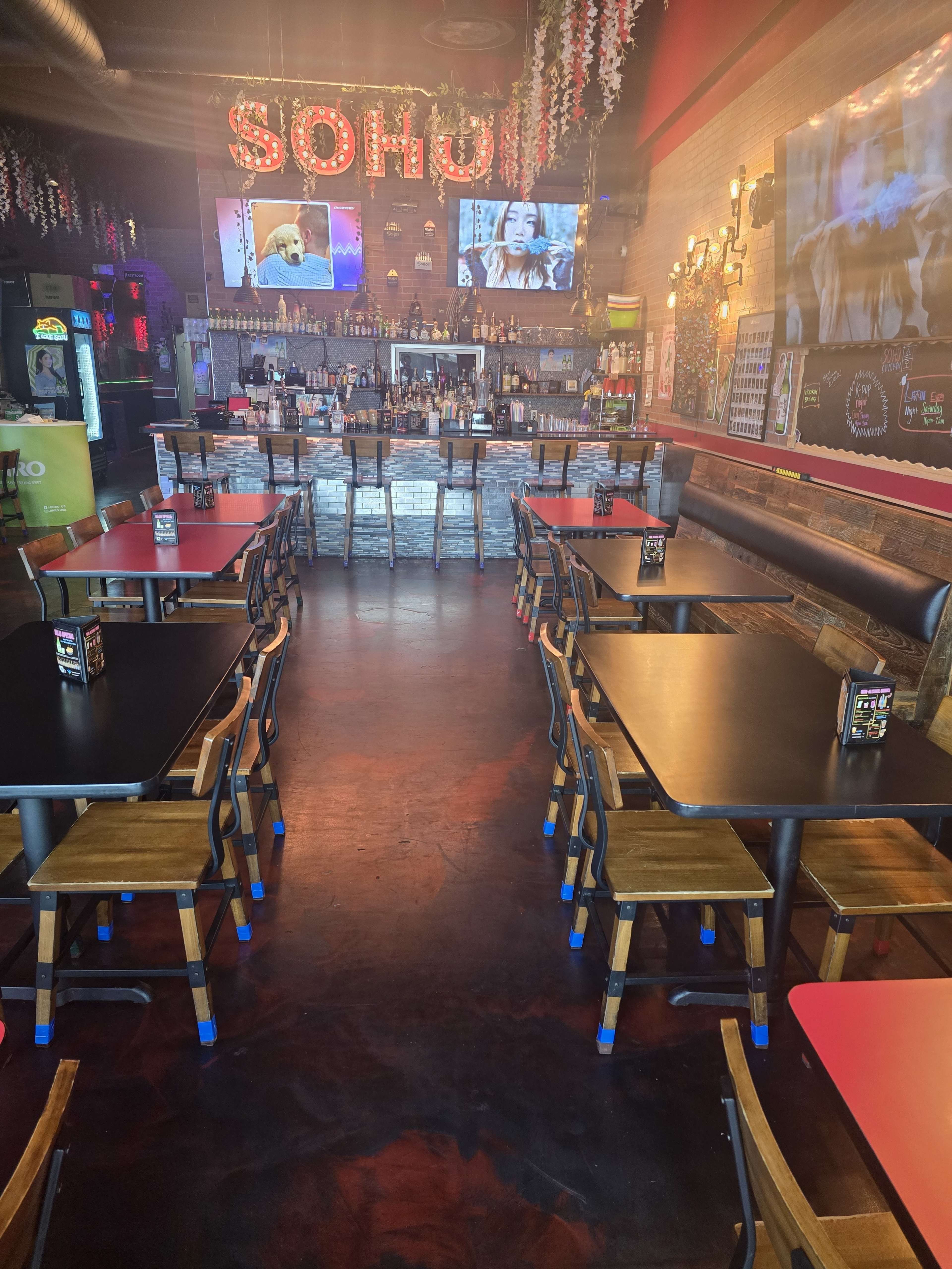 The image shows a restaurant interior with several tables and chairs arranged in front of a bar, illuminated by neon signage and displaying multiple screens on the walls.