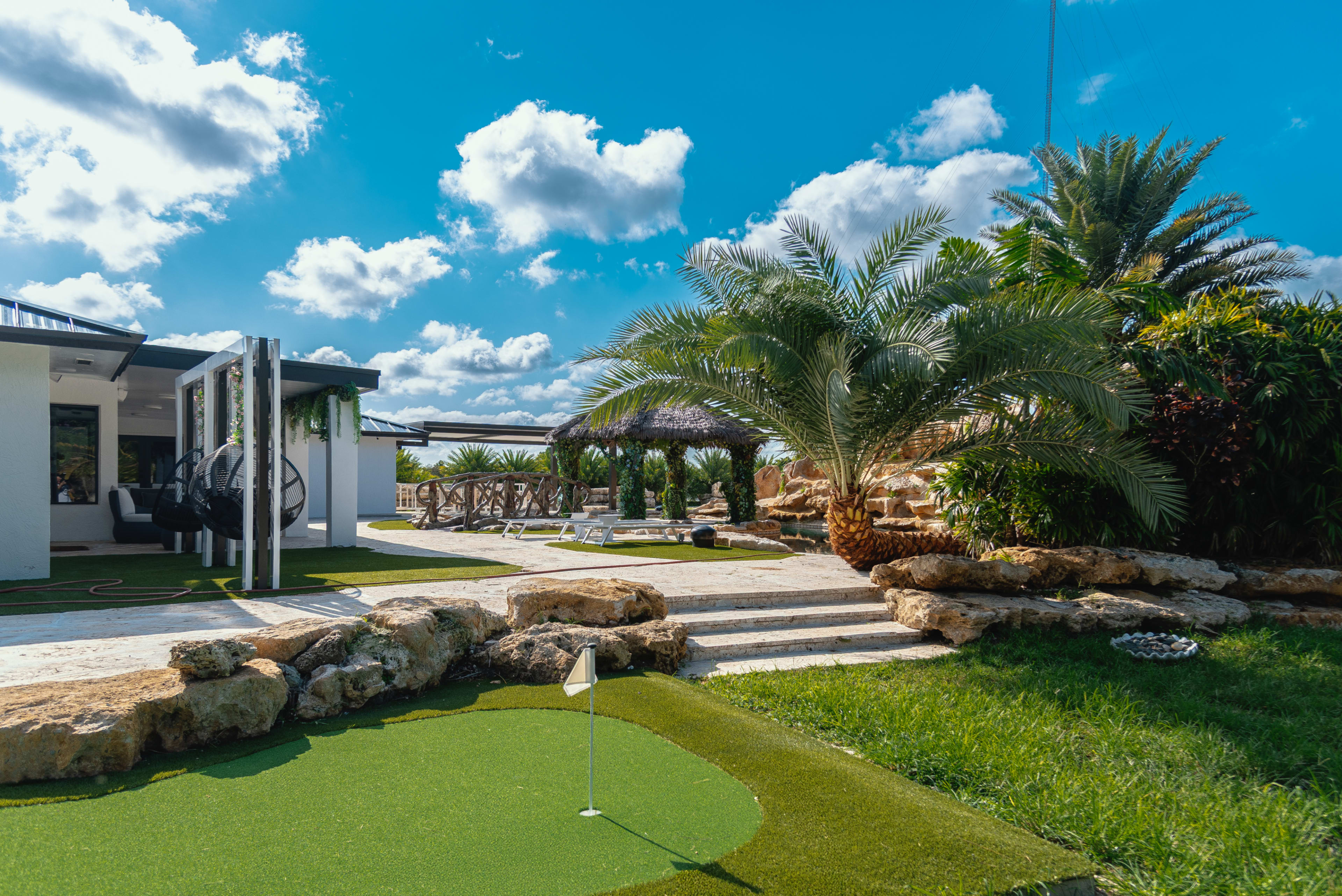 The image shows a landscaped outdoor area featuring a synthetic golf putting green, palm trees, and a modern house with a patio.