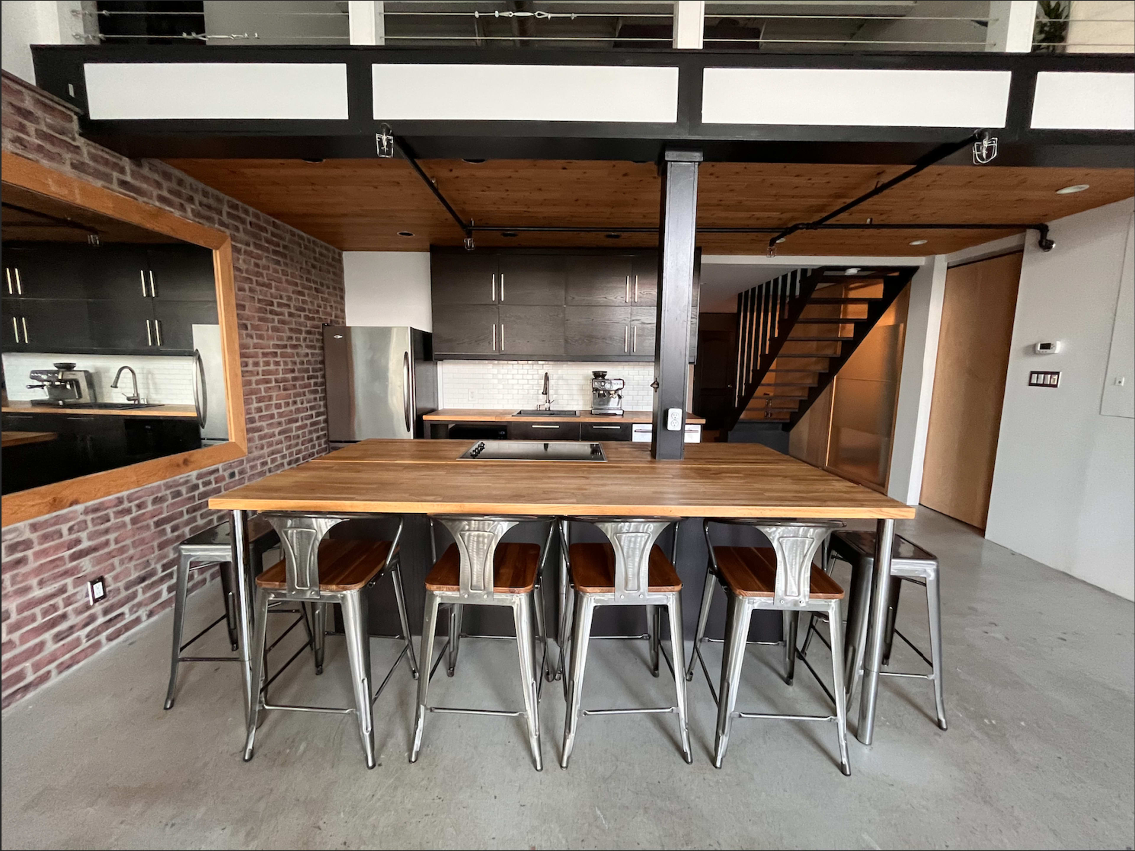 A modern kitchen featuring a wooden island with metallic stools, dark cabinetry, and a staircase in the background.