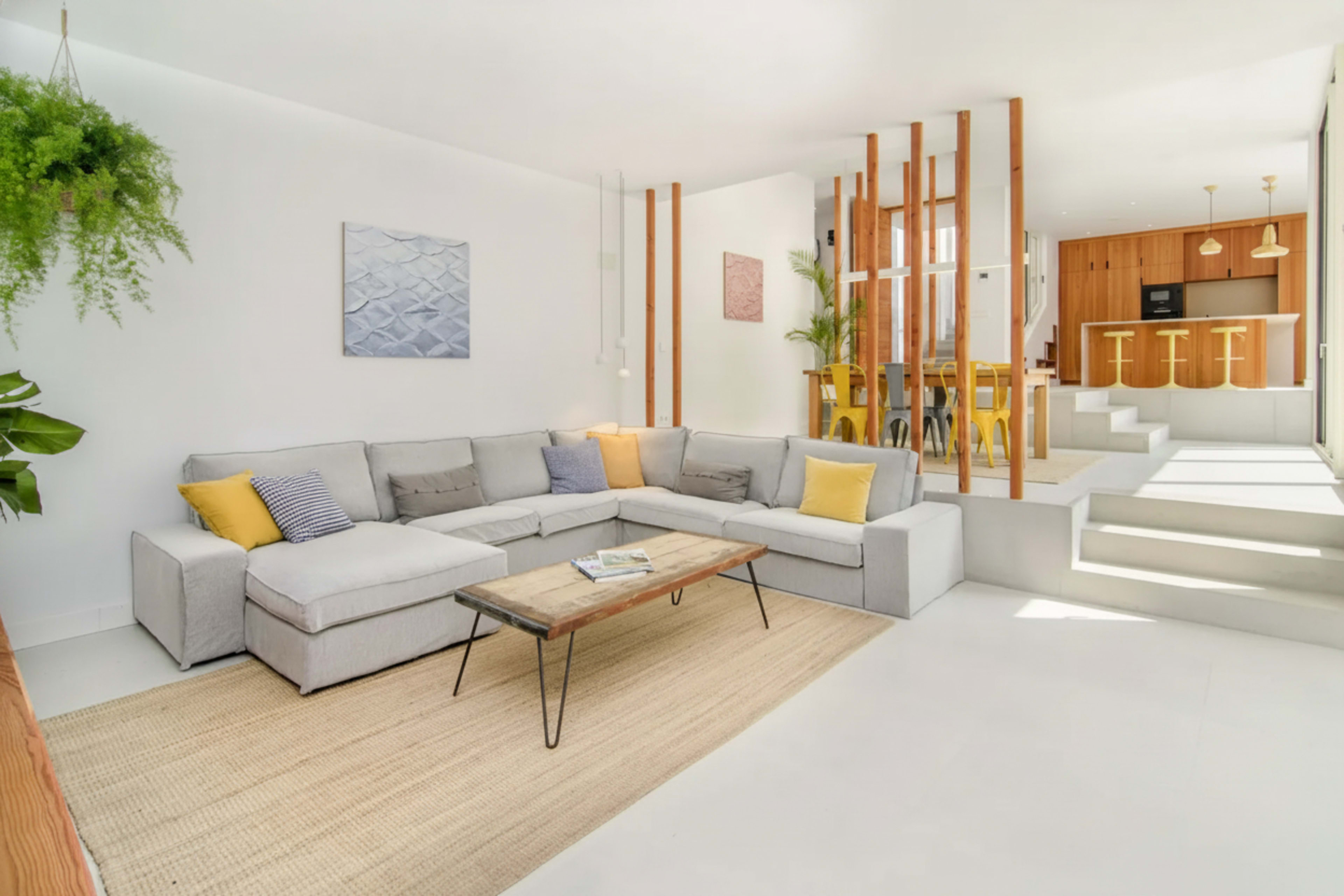 A modern living room features a light gray sectional sofa with yellow and blue accent pillows, a wooden coffee table, and a textured rug, with a kitchen visible in the background.