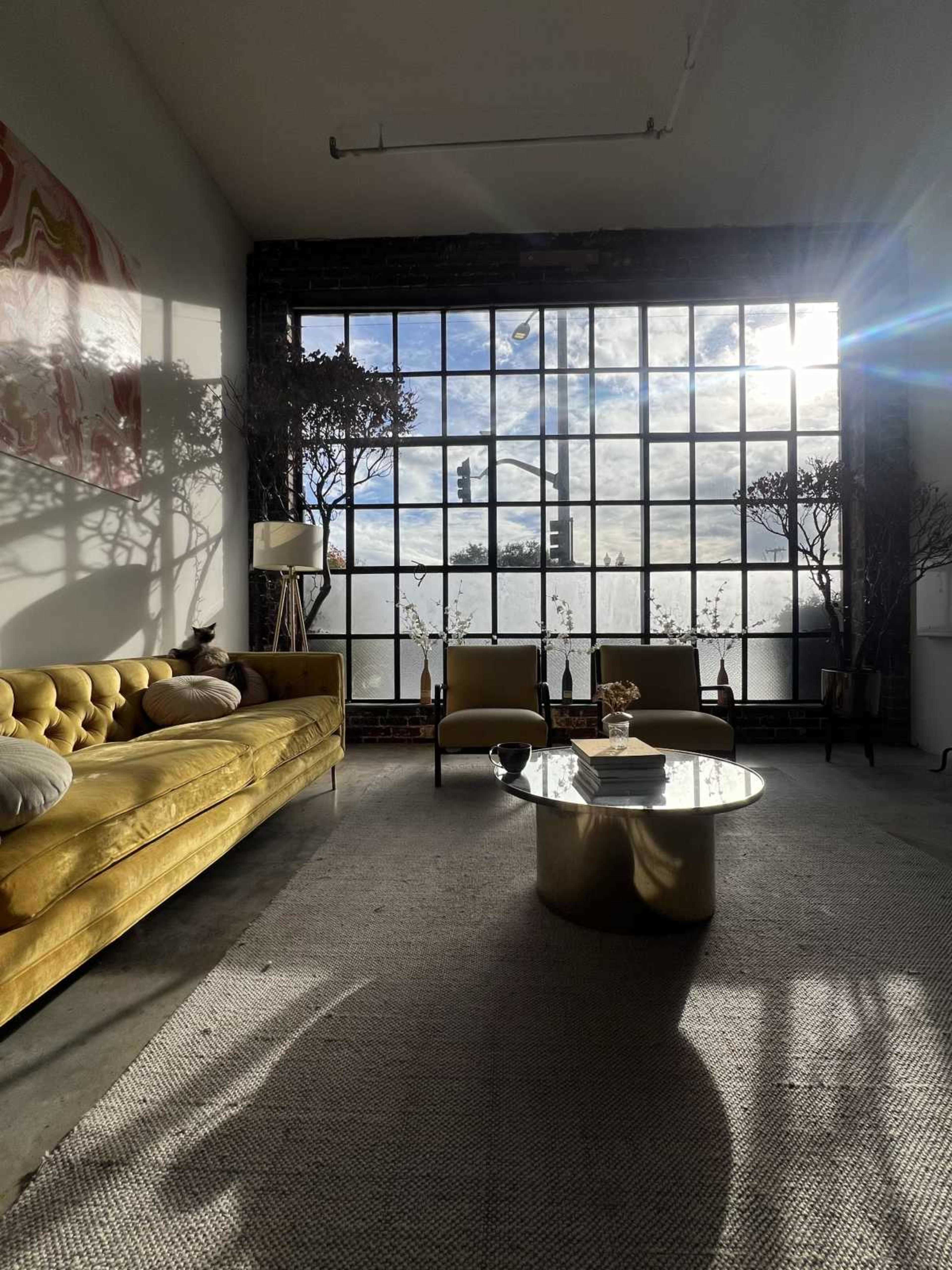 A spacious living room features a yellow sofa, a round coffee table, and large windows that allow natural light to illuminate the space.