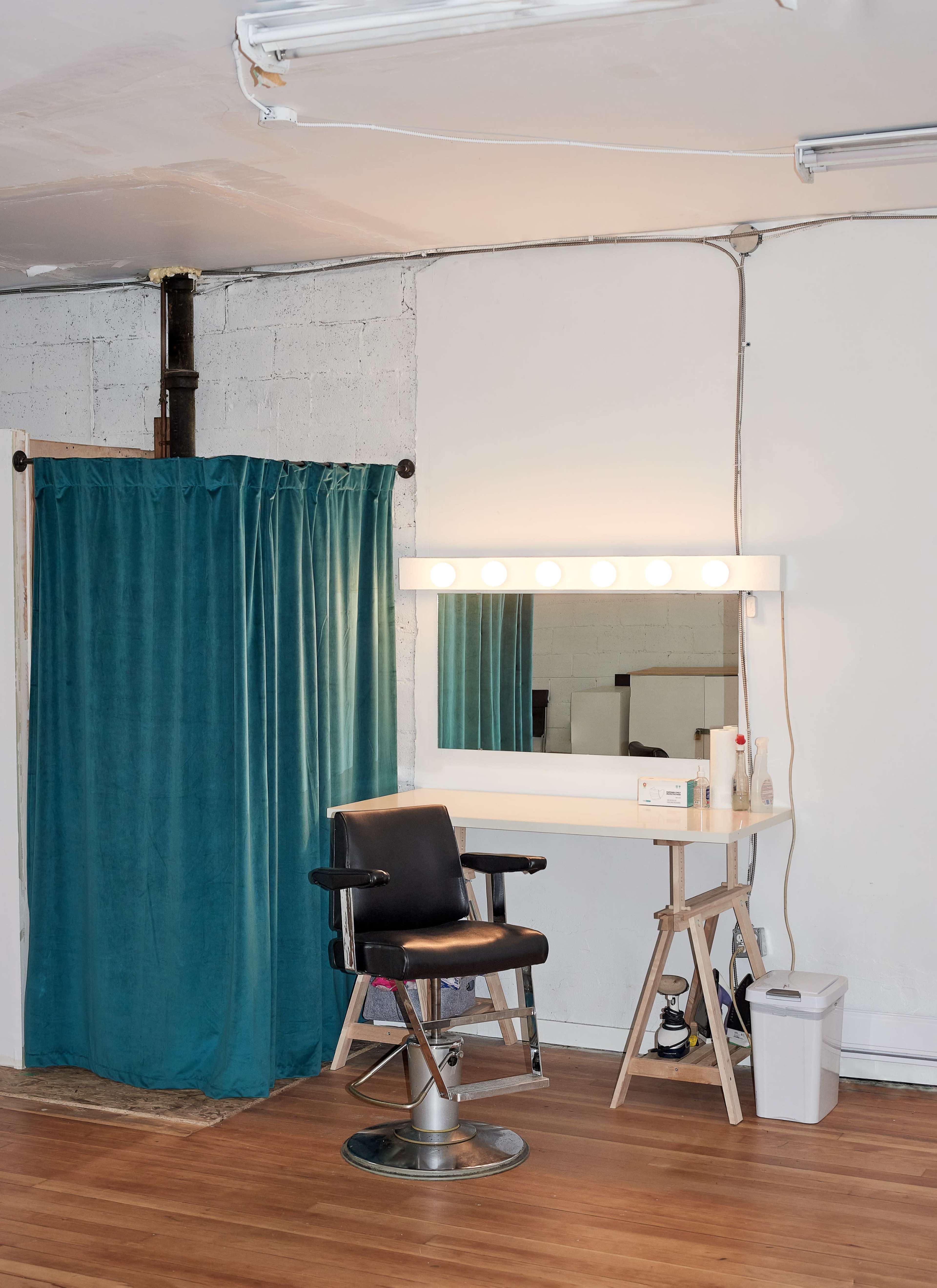 A hair salon setup with a stylist chair in front of a mirror framed by lights, next to a teal curtain.