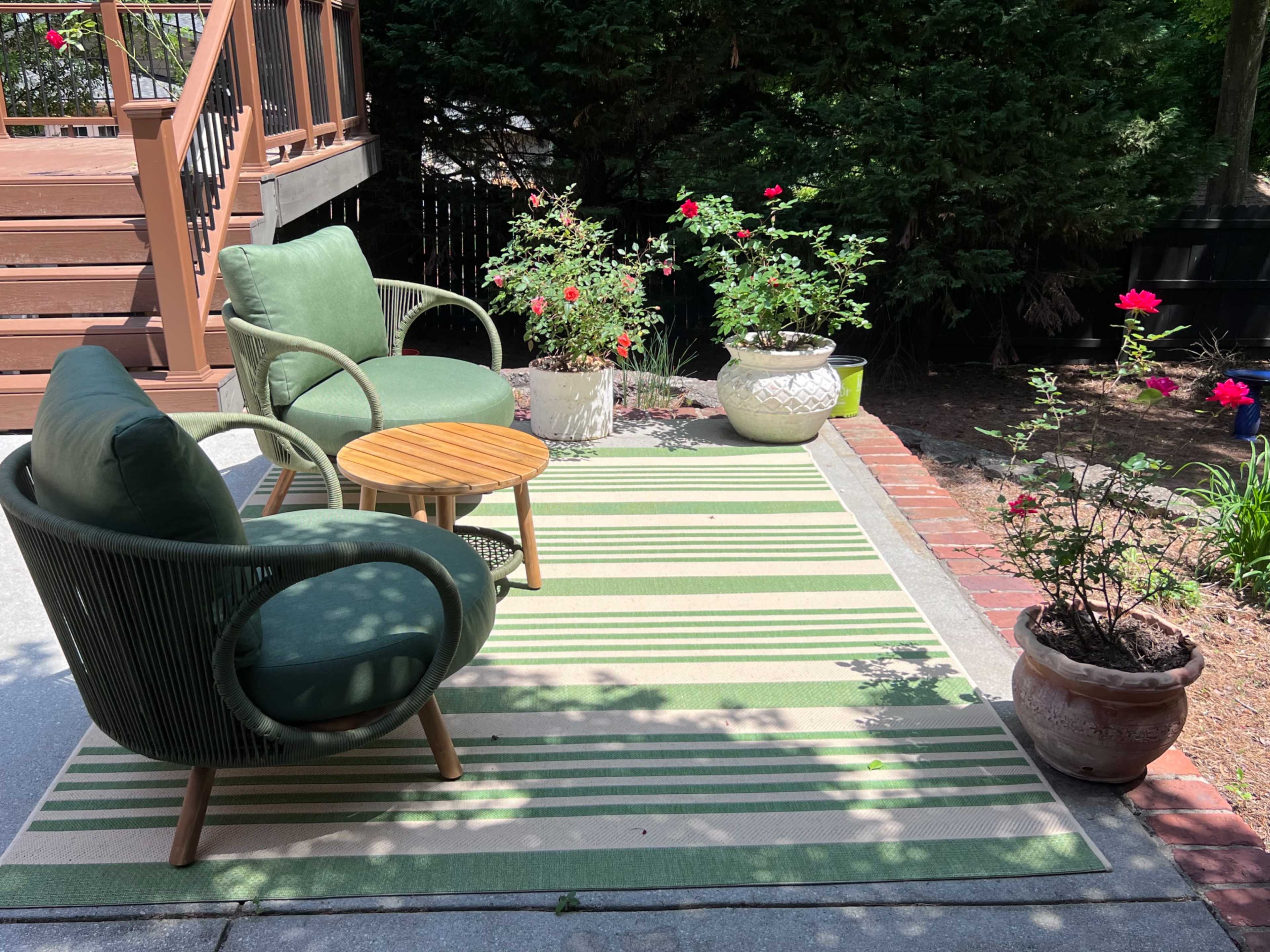 A patio area features two green chairs surrounding a small wooden table, with potted plants and flowers arranged along the edges.