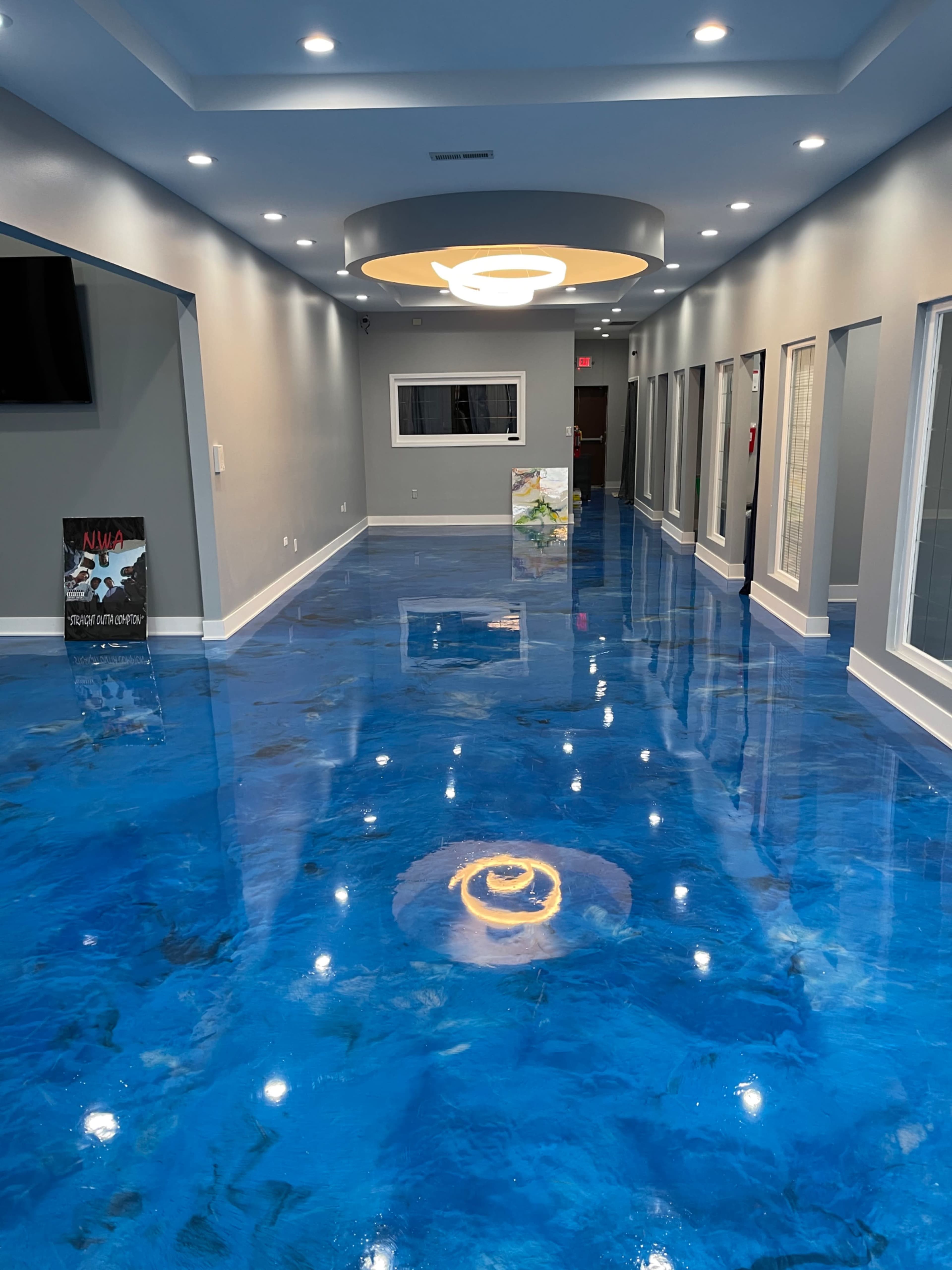 The image shows a modern hallway with a shiny blue epoxy floor, illuminated by overhead lighting and lined with glass-paneled doors.