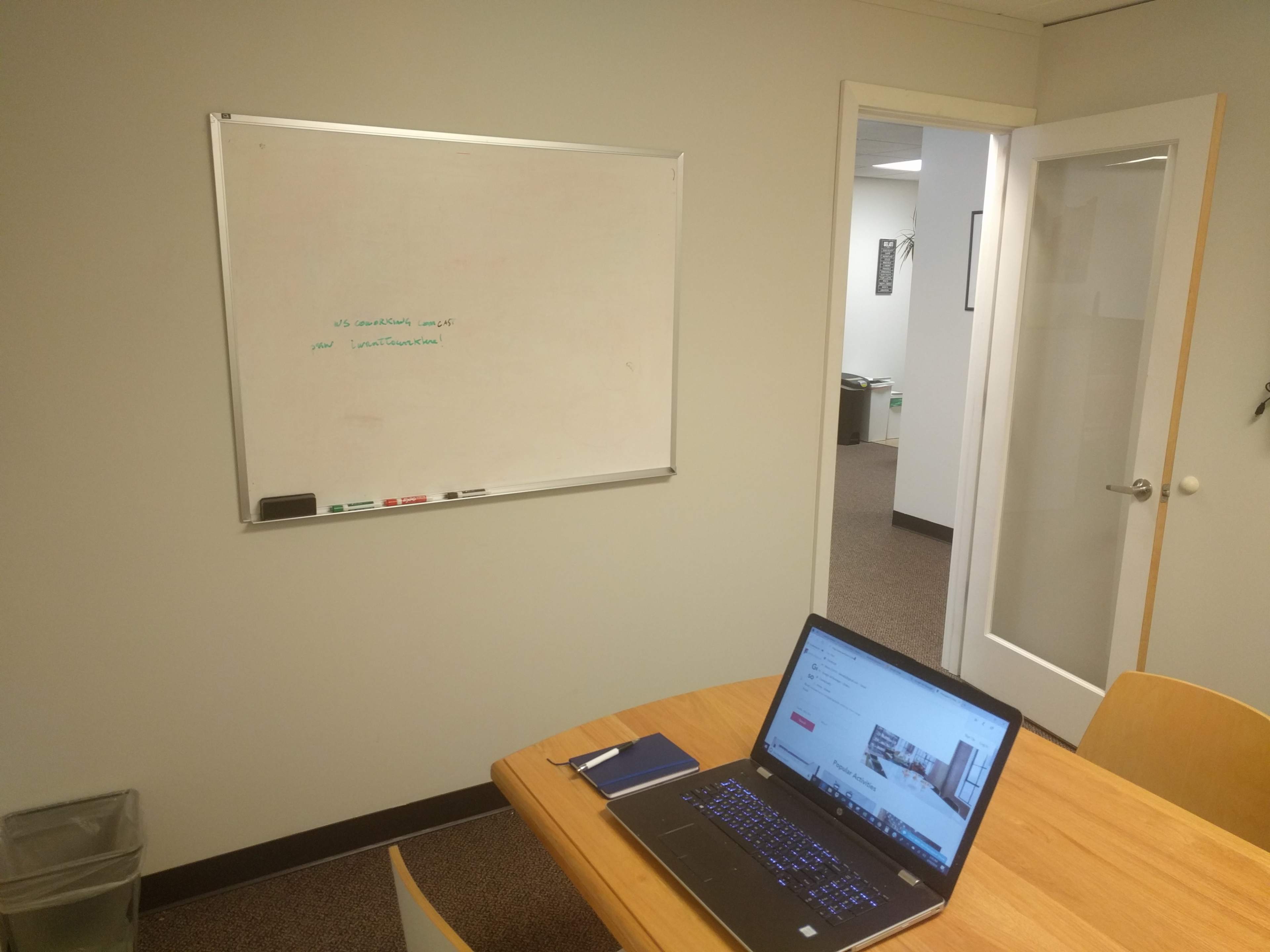 A laptop sits on a wooden table next to a notebook, while a whiteboard is mounted on the wall and a door leads to another room.
