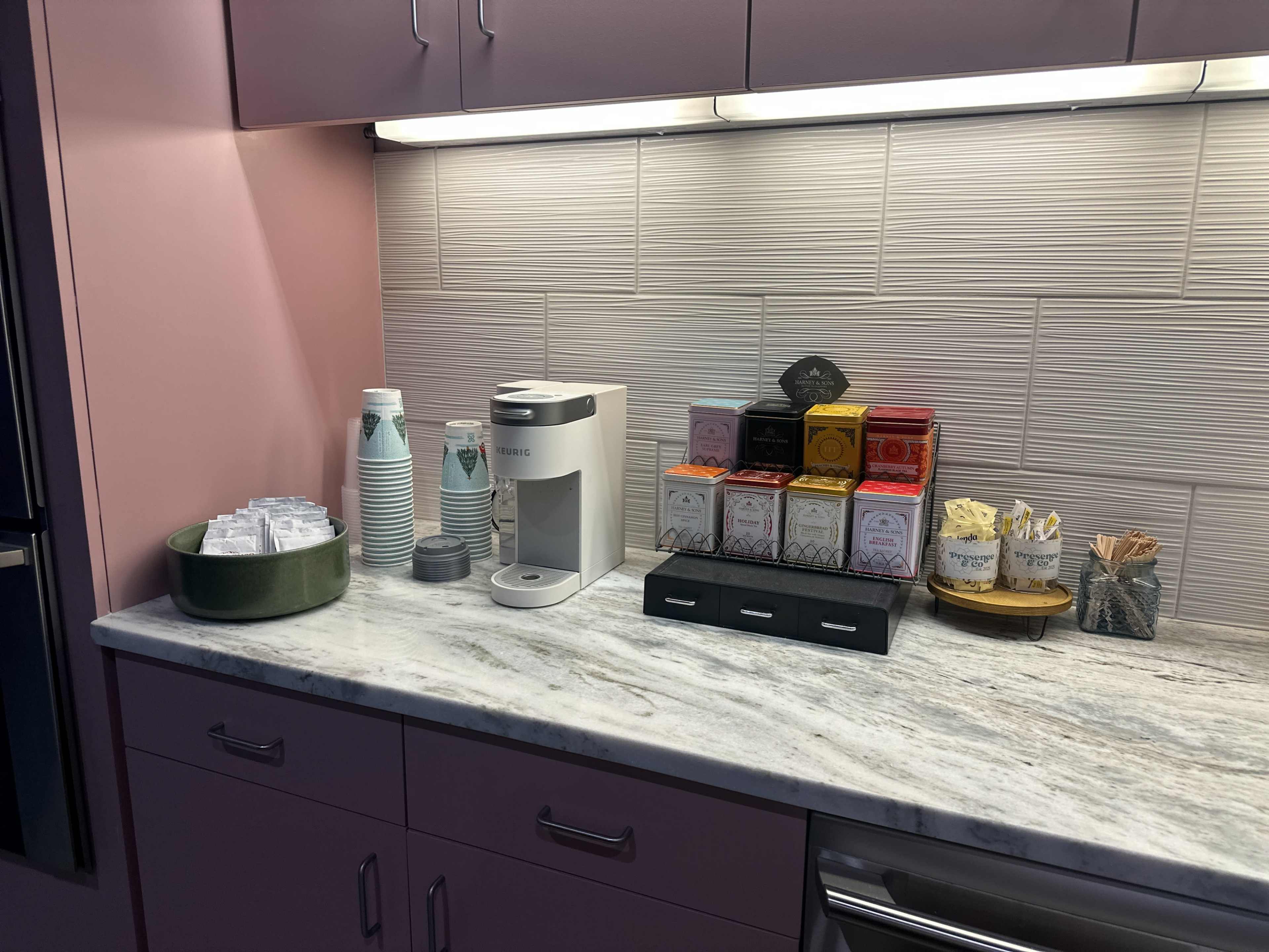 The image shows a kitchen countertop with a coffee maker, various tea boxes, a bowl of sweeteners, disposable cups, and a decorative tray with stirring sticks.