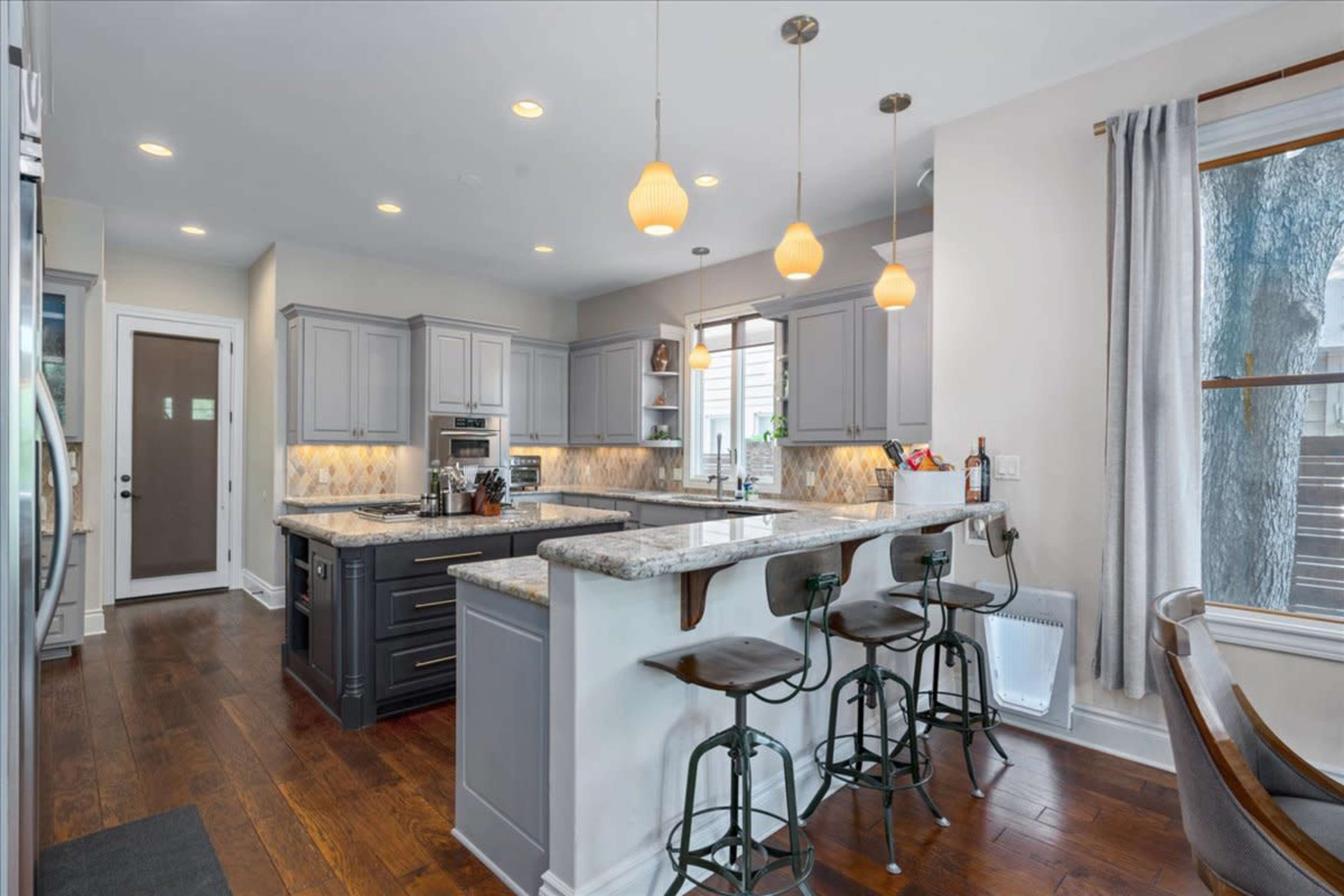The kitchen features gray cabinetry, a large island with bar stools, and pendant lighting over a granite countertop.